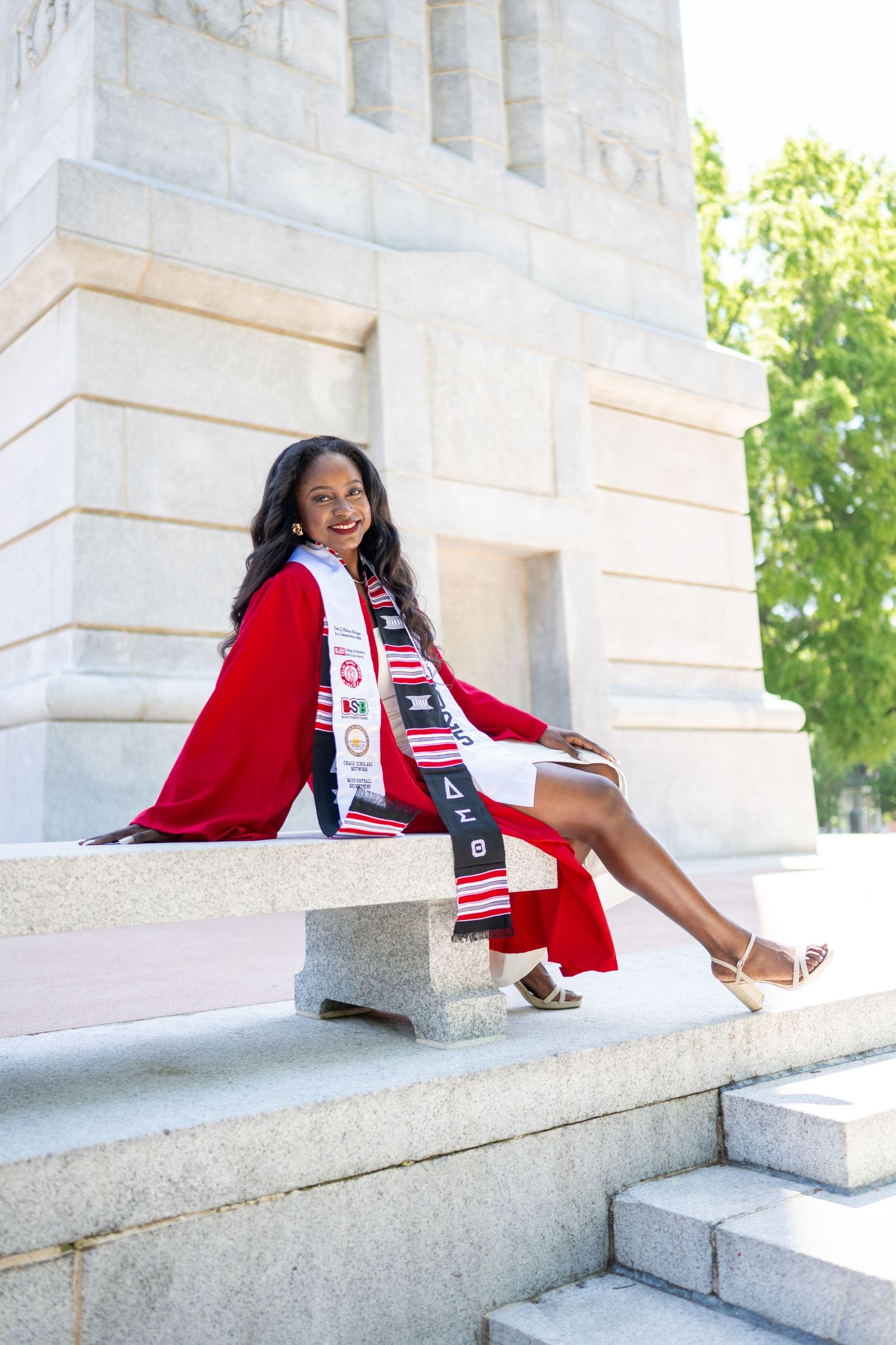Graduate in red academic regalia poses on stone steps during graduation photo session.