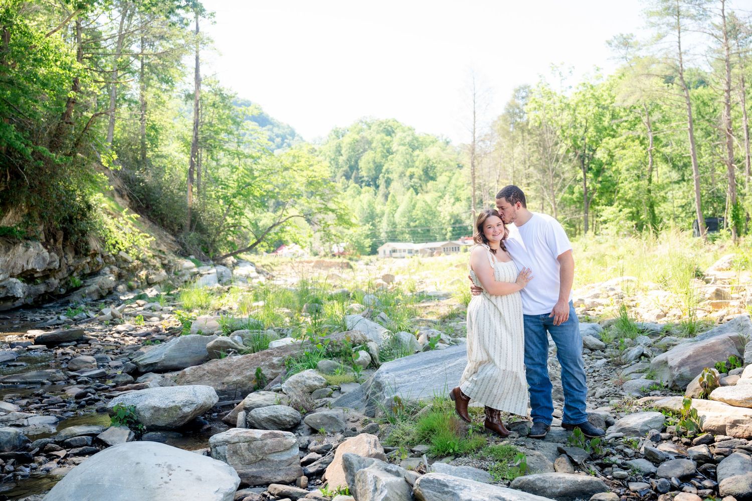 Couple embracing romantically by a rocky mountain stream surrounded by lush green trees in a scenic outdoor setting.