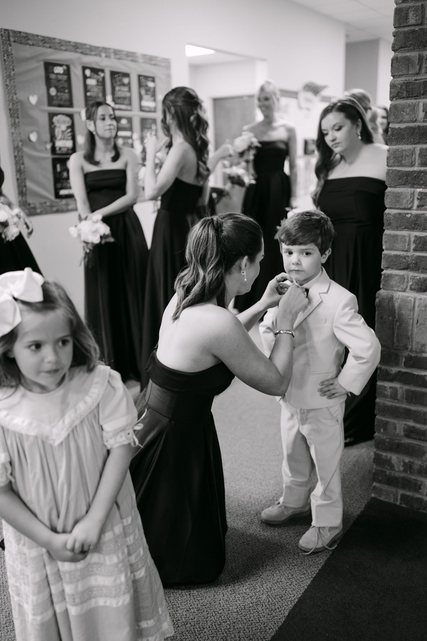 Black and white wedding photo of bridesmaids in black dresses helping young ring bearer adjust his tie.