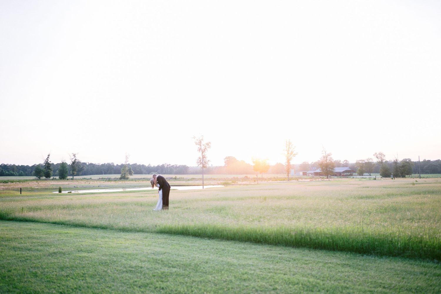 Couple embracing in a vast green field at golden sunset, with trees and water in the background.