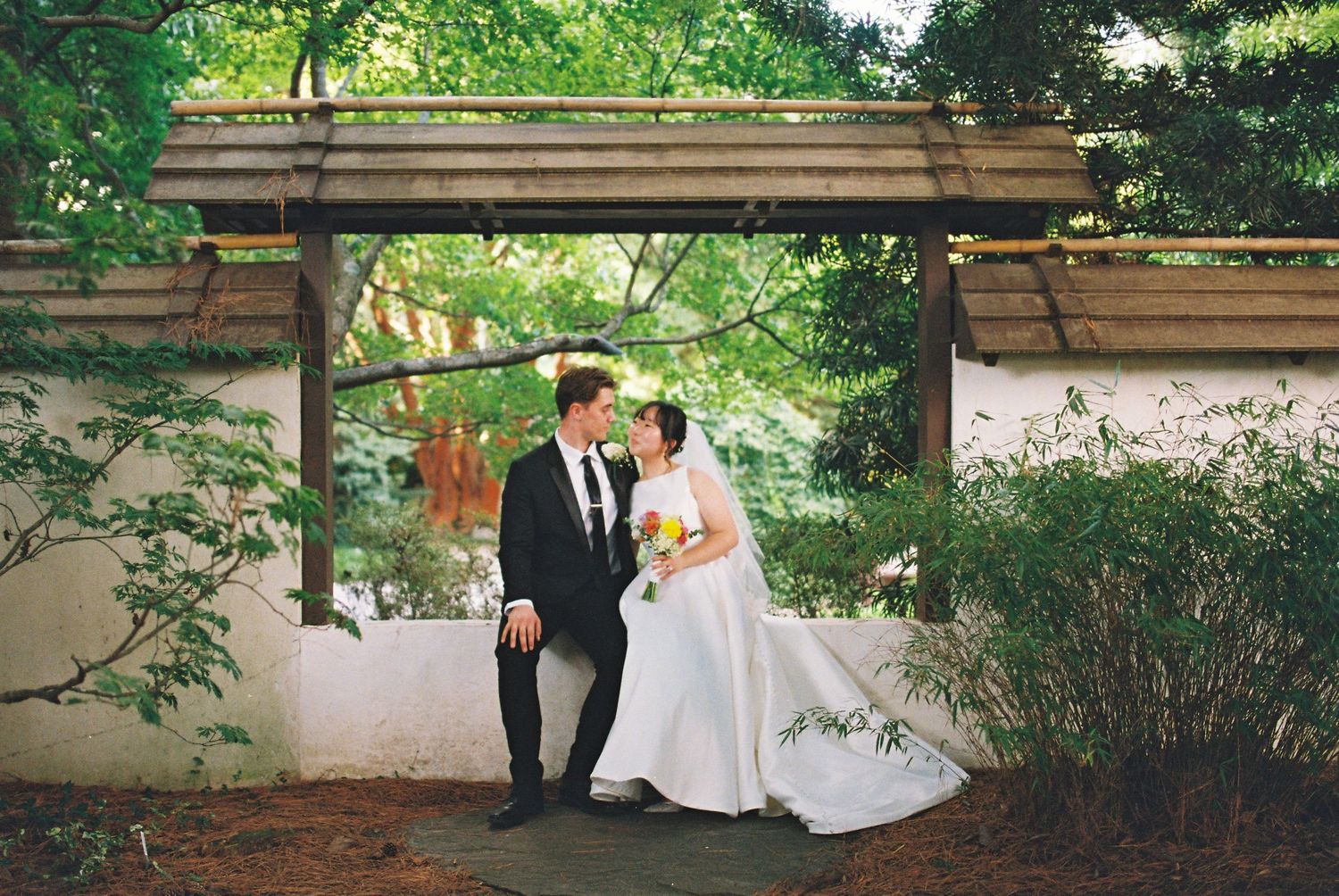 Bride and groom share a romantic moment under a wooden pergola surrounded by lush green trees in a garden wedding.