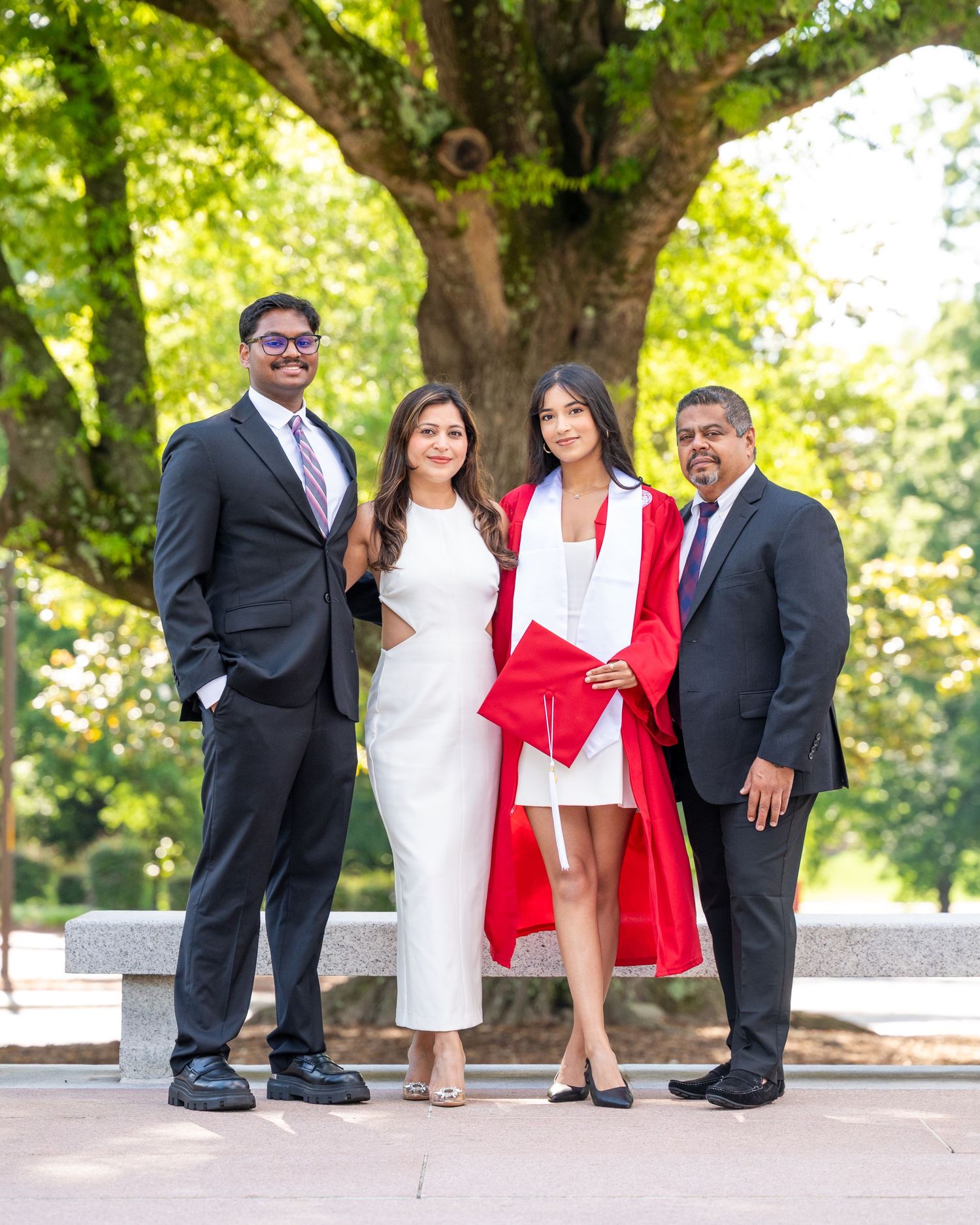 A group in formal attire poses together on stone steps under green trees in a park setting.
