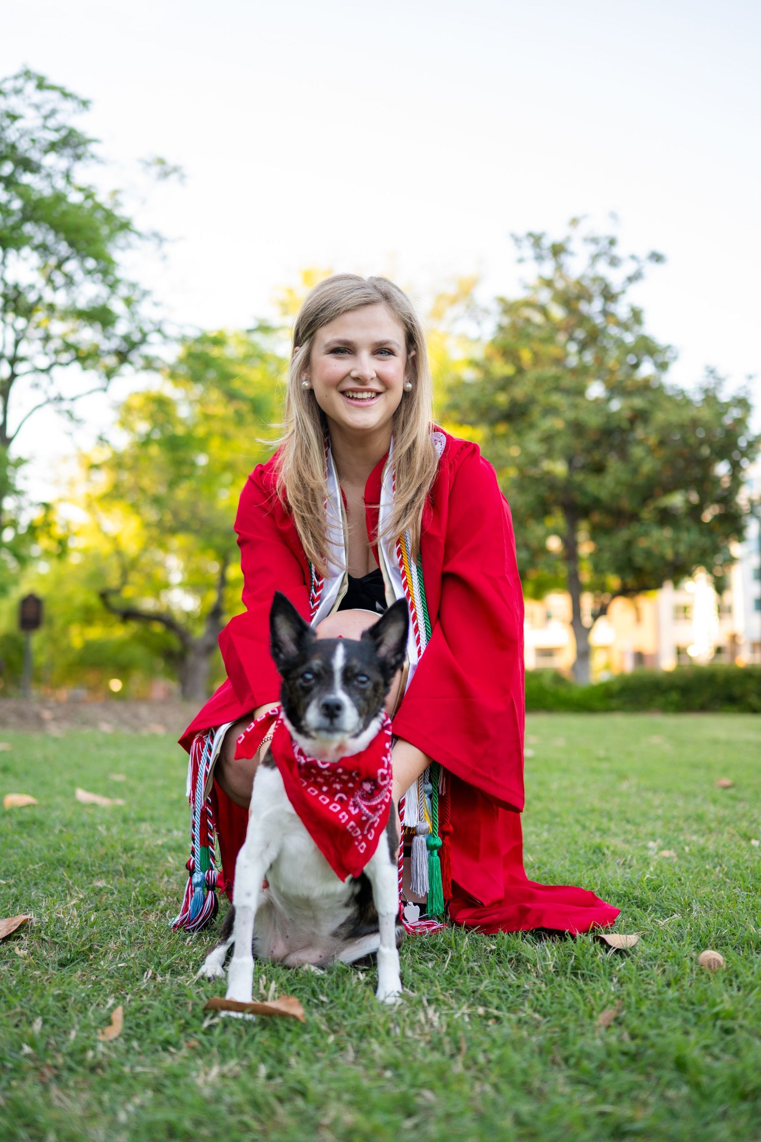 A small black and white dog wearing a red bandana sits on grass next to someone in a bright red coat on a sunny day.