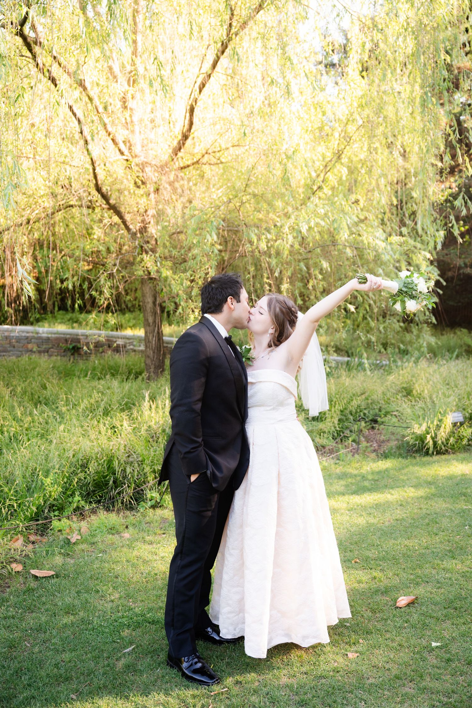 Bride and groom share a romantic kiss under a weeping willow tree on a sunny day in a lush green garden.