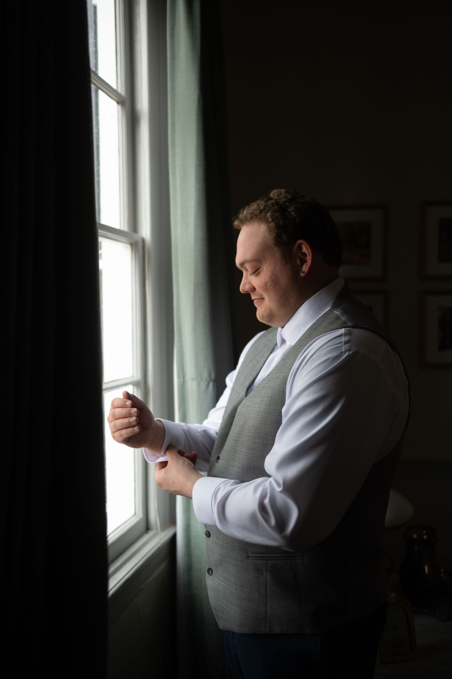 Groom in gray vest fastening cufflinks by a bright window, preparing for his wedding day.
