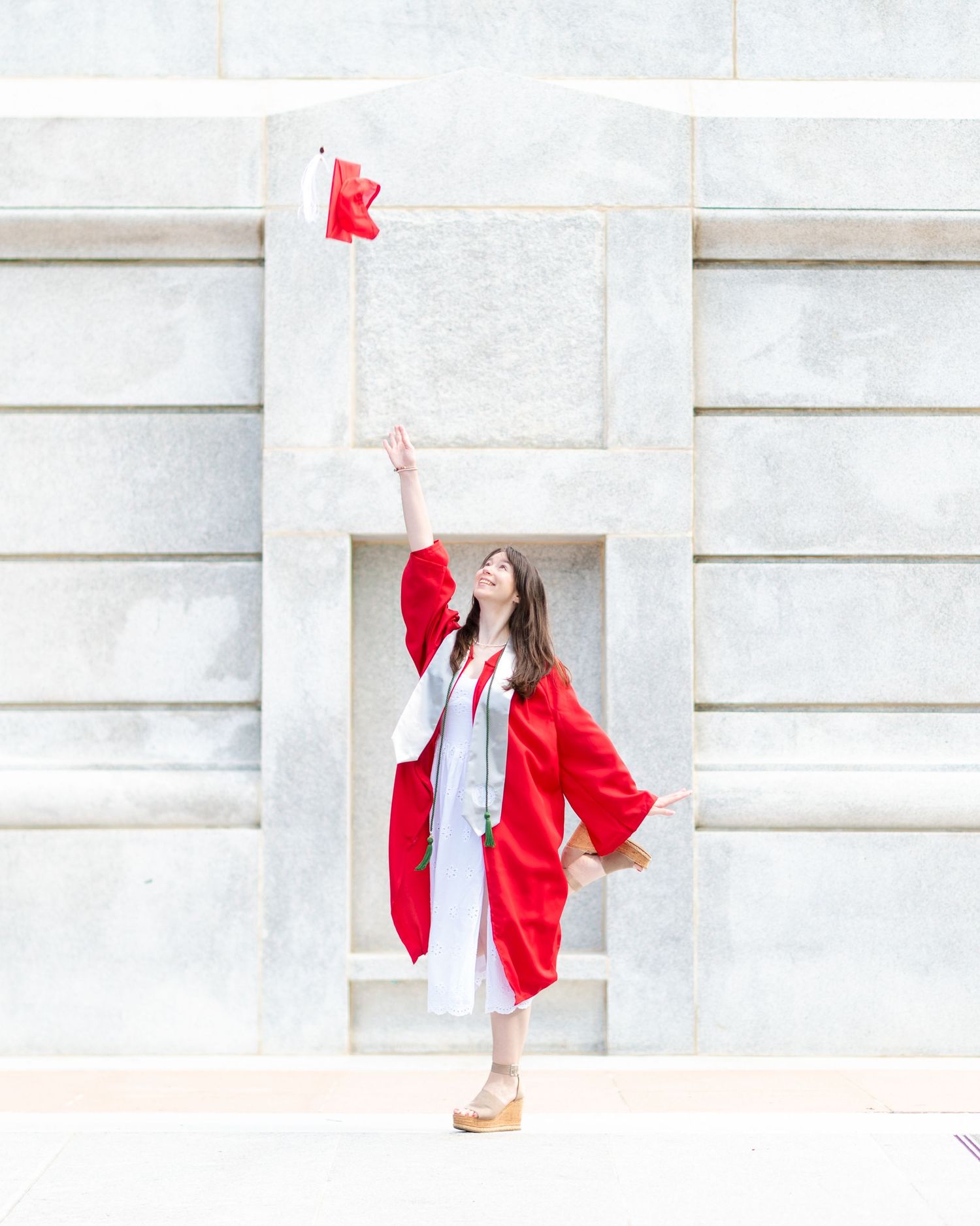 Graduate in white dress and red graduation gown jumps joyfully while holding diploma against light stone wall.