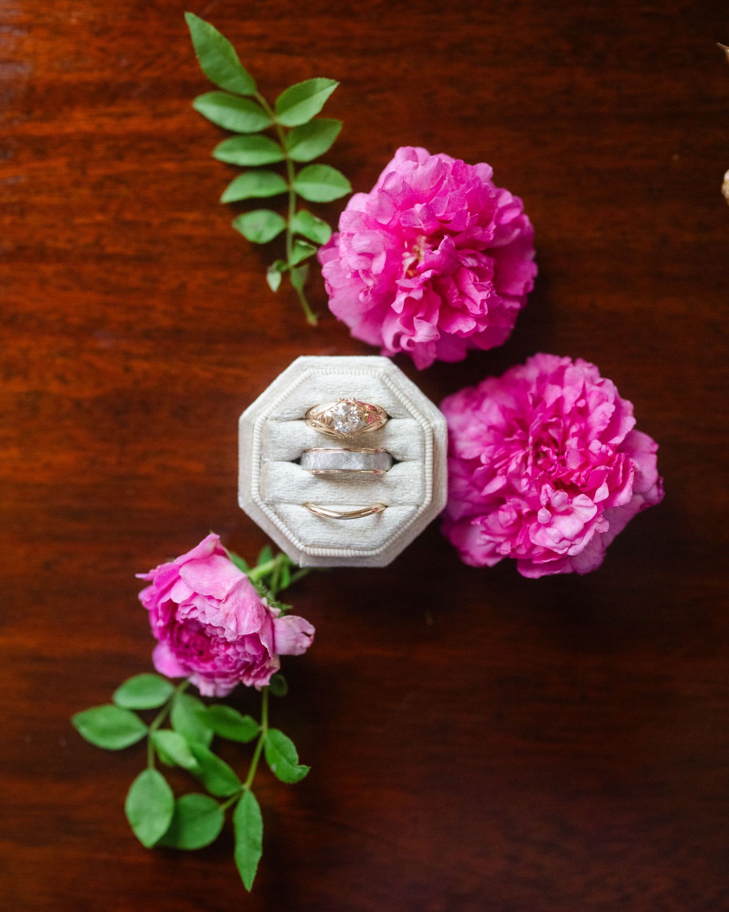An engagement ring in a white octagonal box surrounded by vibrant pink peonies and green leaves on a wooden surface.