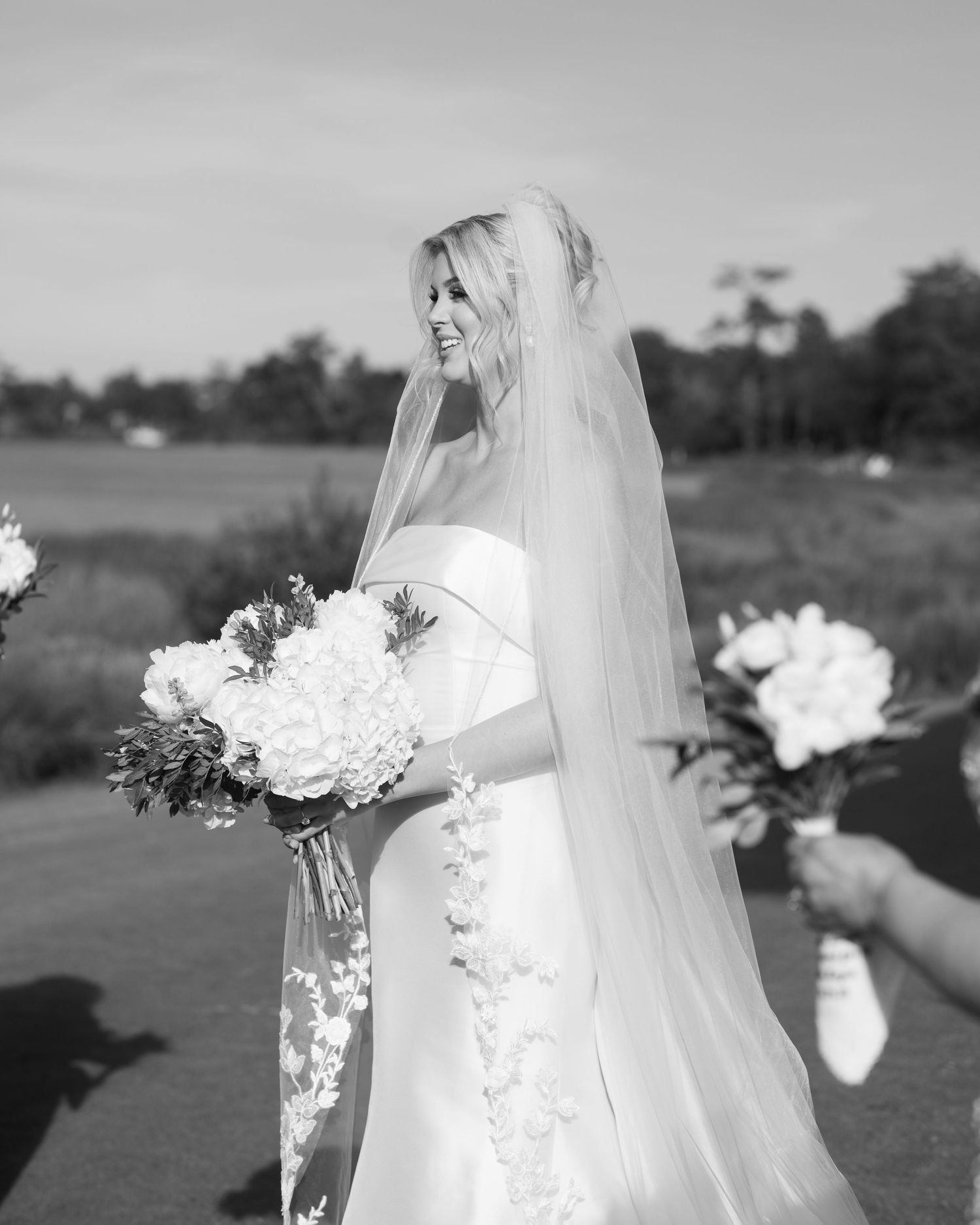 Black and white photo of a bride in a strapless gown and long veil holding a white floral bouquet outdoors.