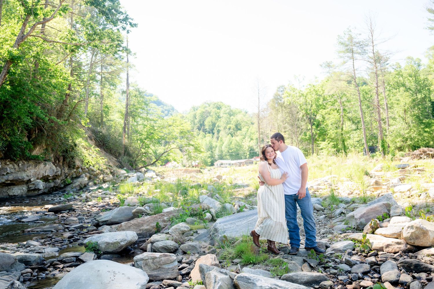 Couple embracing by a rocky creek surrounded by lush green trees on a sunny day.