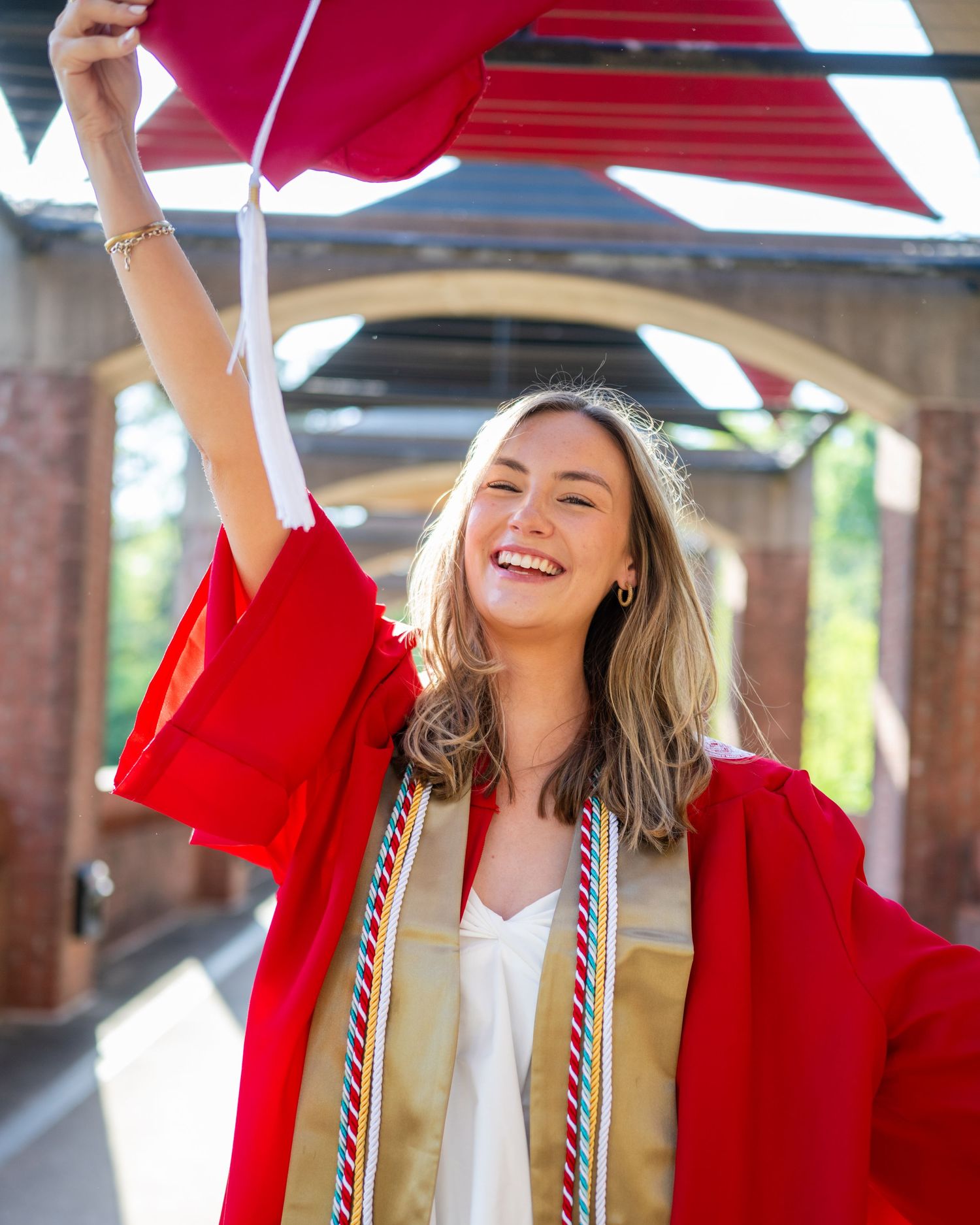 Graduate in red academic regalia and graduation cords smiles joyfully while celebrating under red banners outdoors.