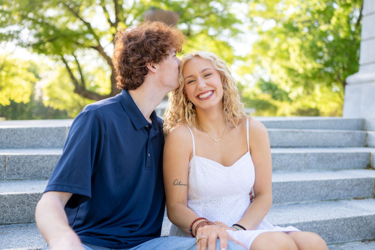 A couple sits together on outdoor concrete steps during a sunny day, sharing a joyful moment.