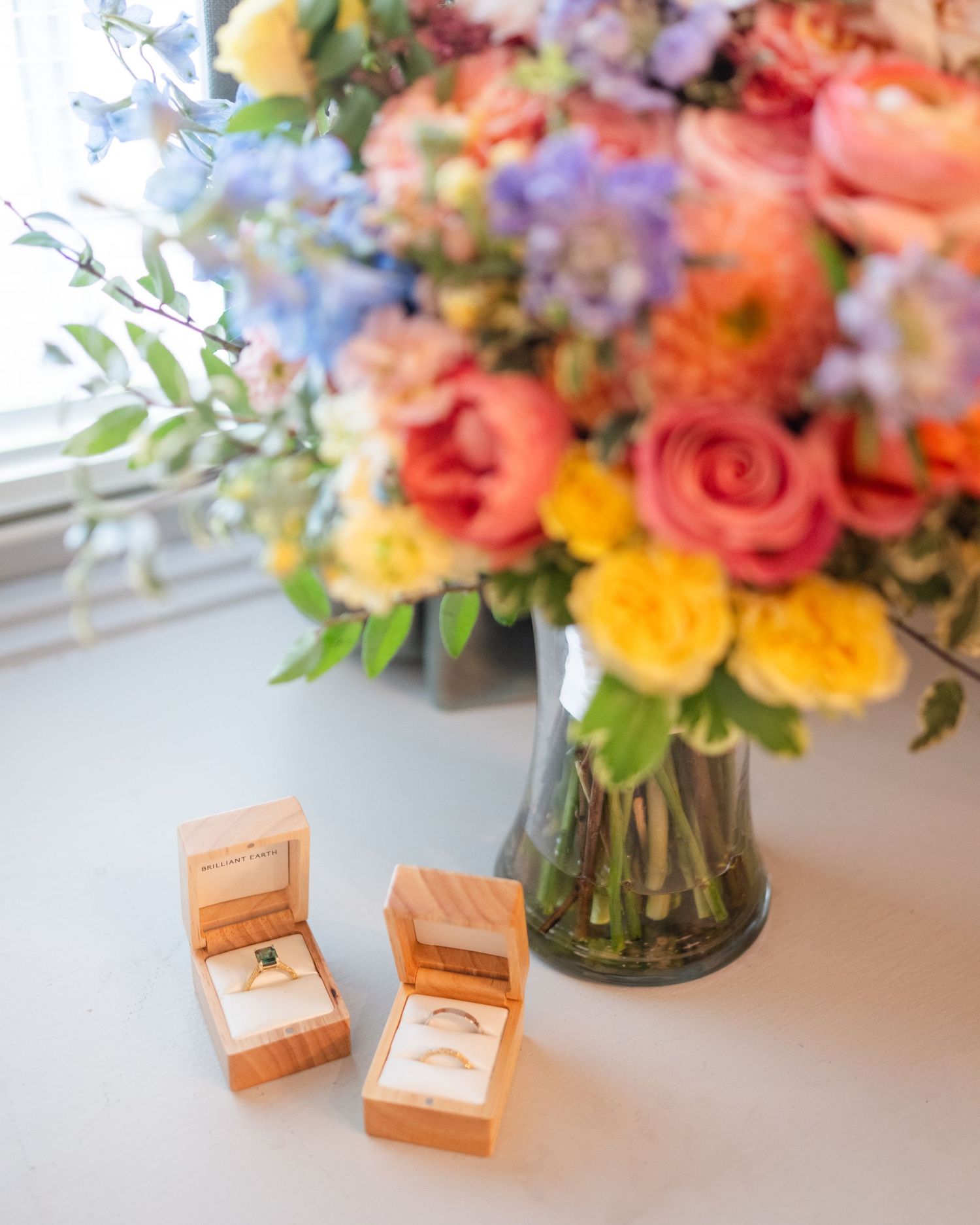 Colorful floral centerpiece with roses and delicate blooms sits next to wooden ring boxes on a white surface.