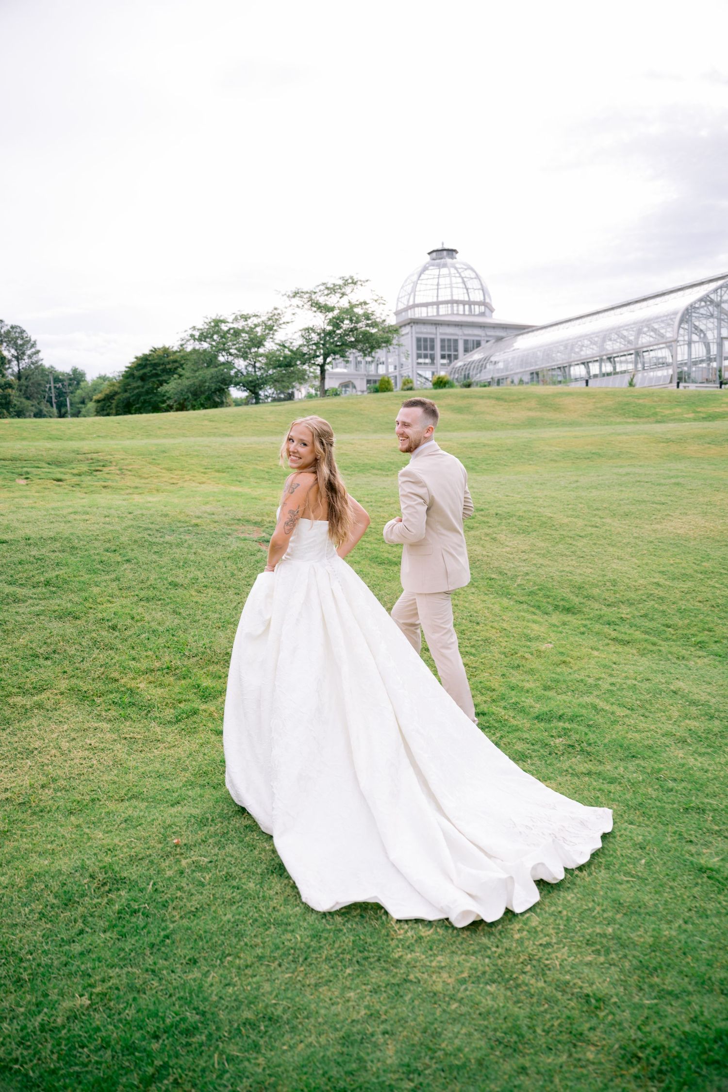 Bride in white ball gown and groom in tan suit walk hand in hand on lush green lawn with greenhouse backdrop.