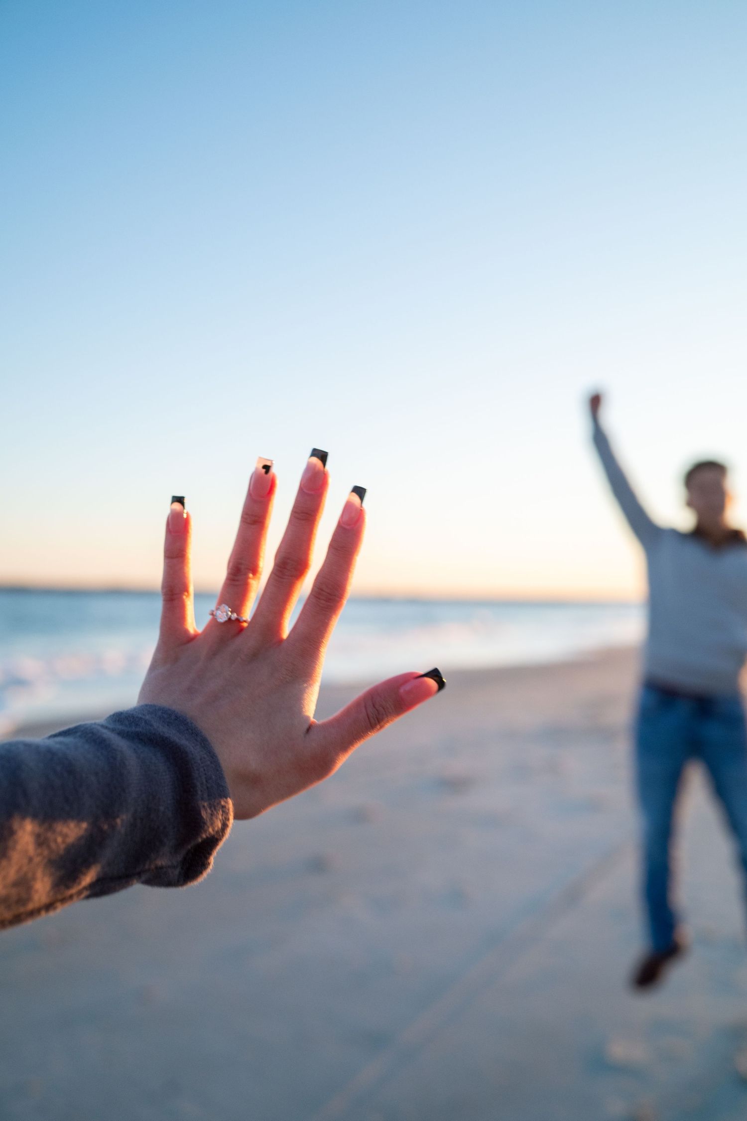 Hand with black nail polish and engagement ring reaches toward silhouetted figure on beach at sunset.