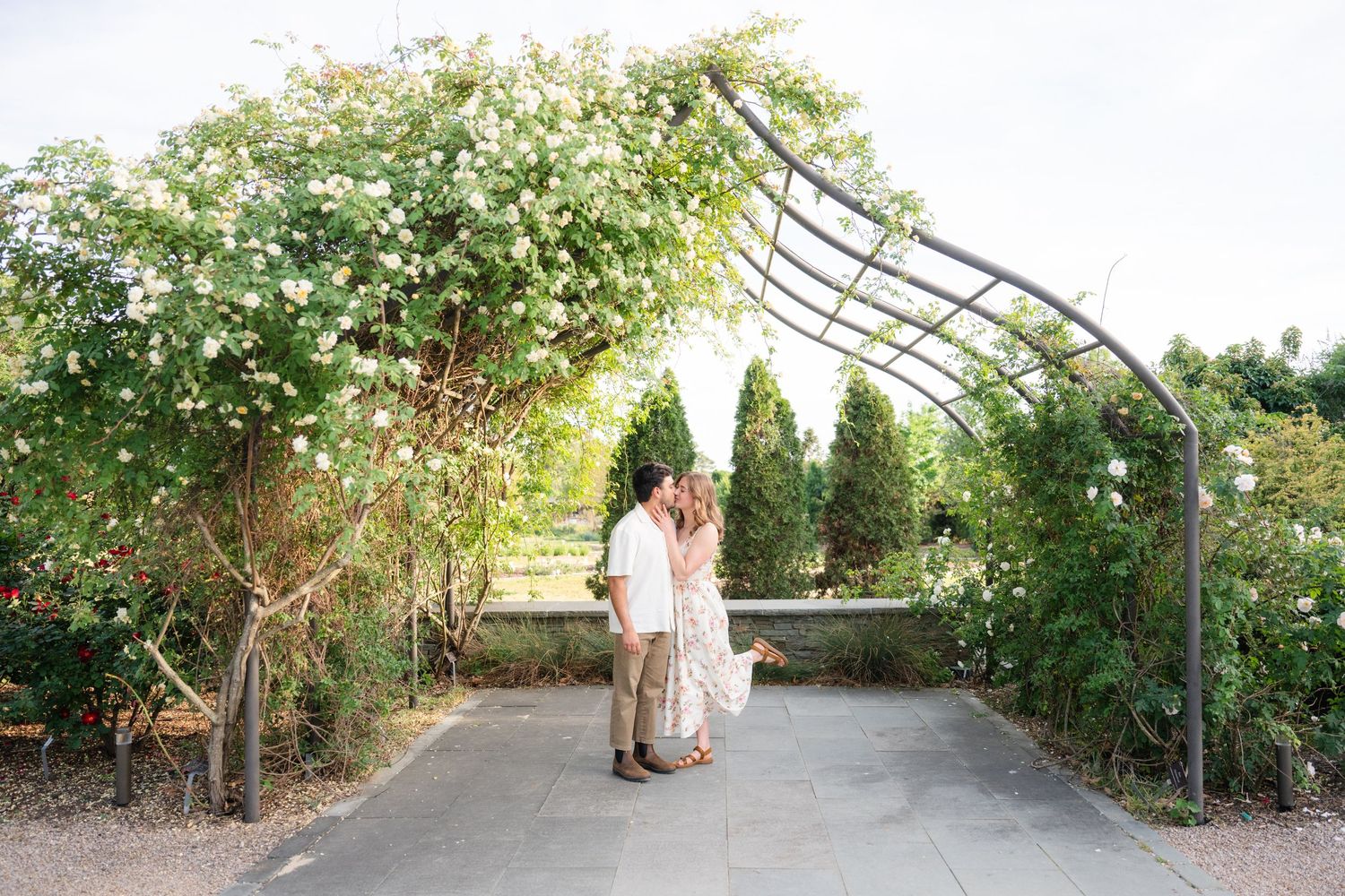 Couple sharing a romantic kiss under a floral garden arch surrounded by lush greenery and blooming roses.