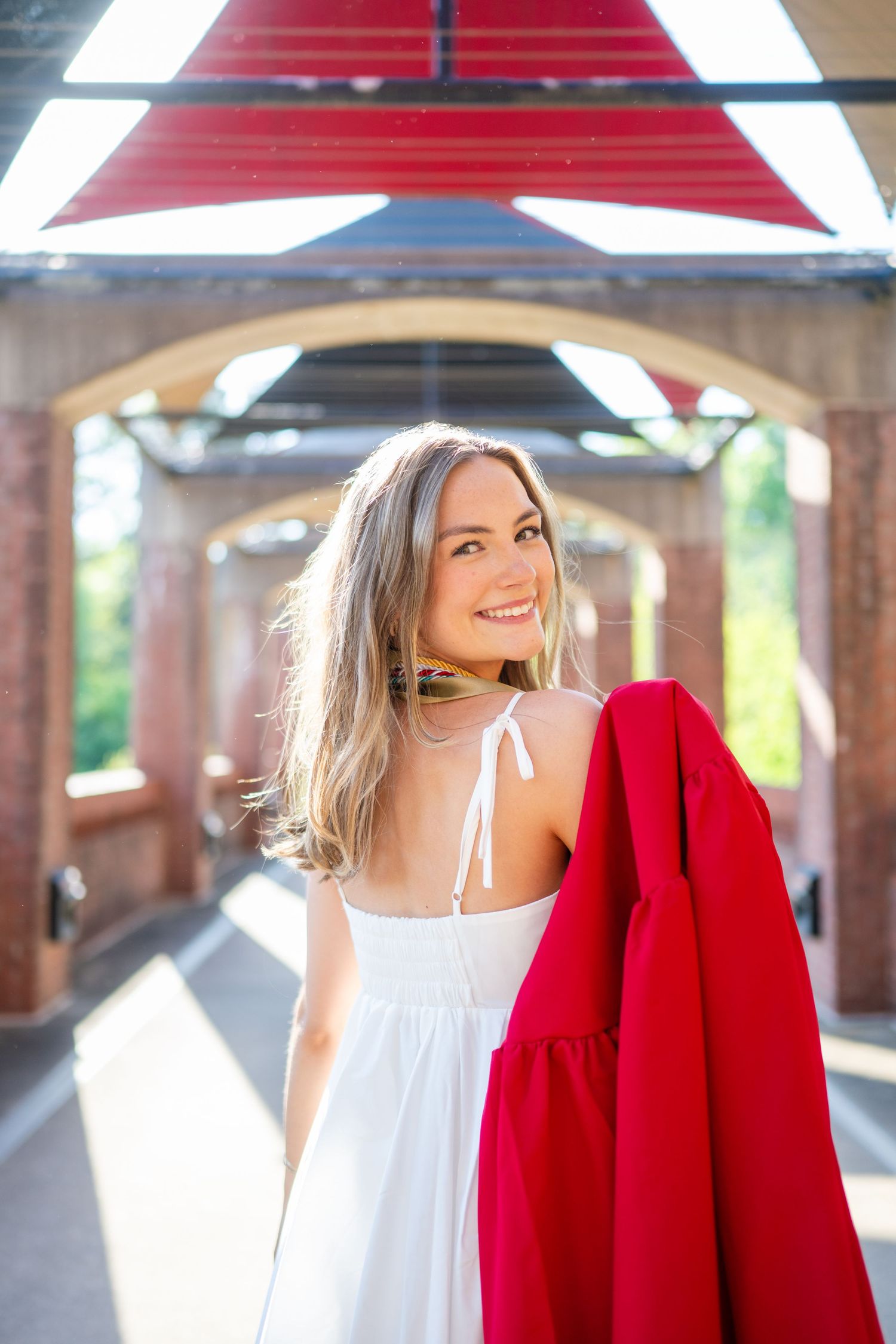 A woman in a white summer dress holds a red coat while standing in a covered walkway on a sunny day.