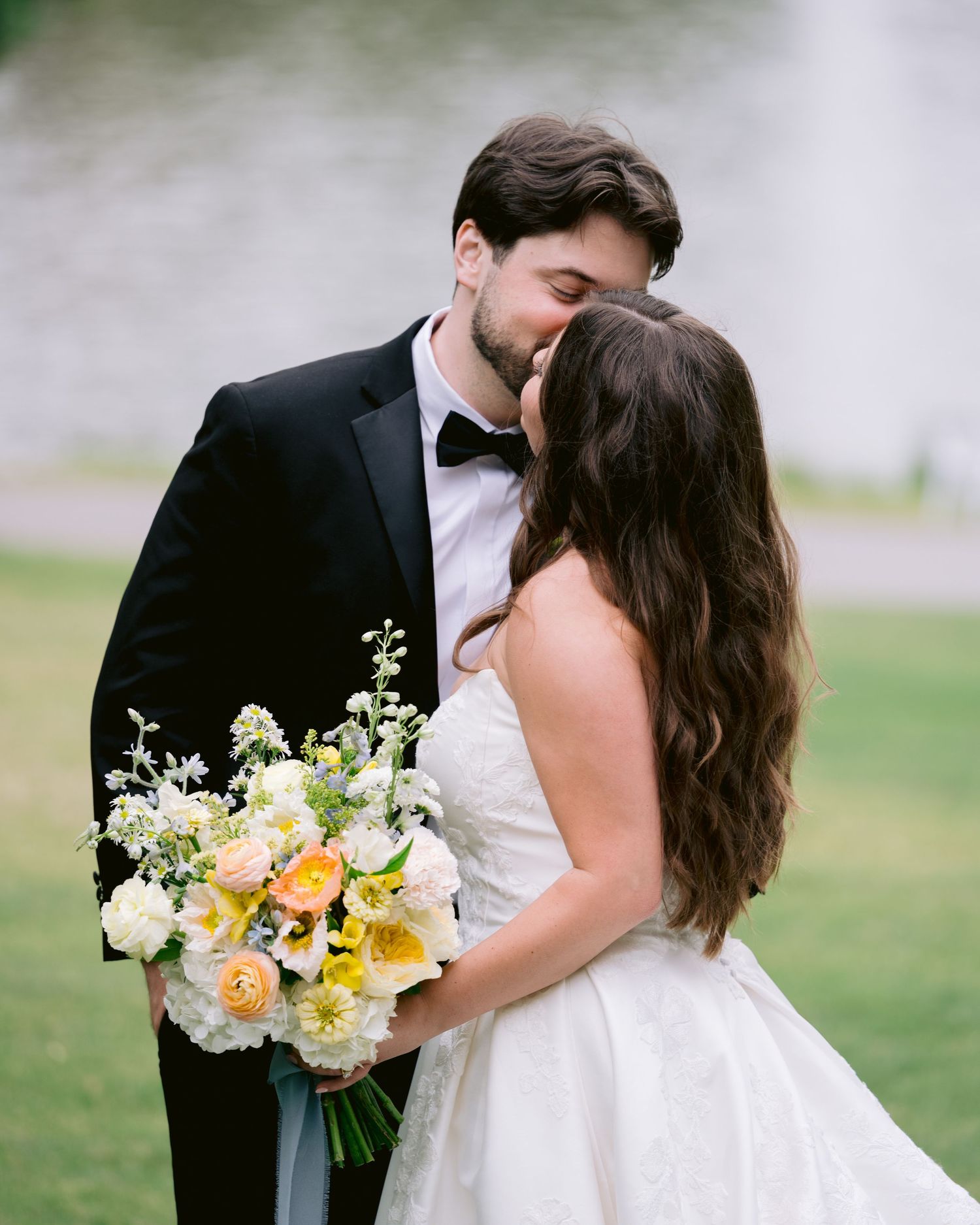 A bride and groom share a romantic kiss, she holds a yellow and white bouquet, he wears a black tuxedo, outdoors.