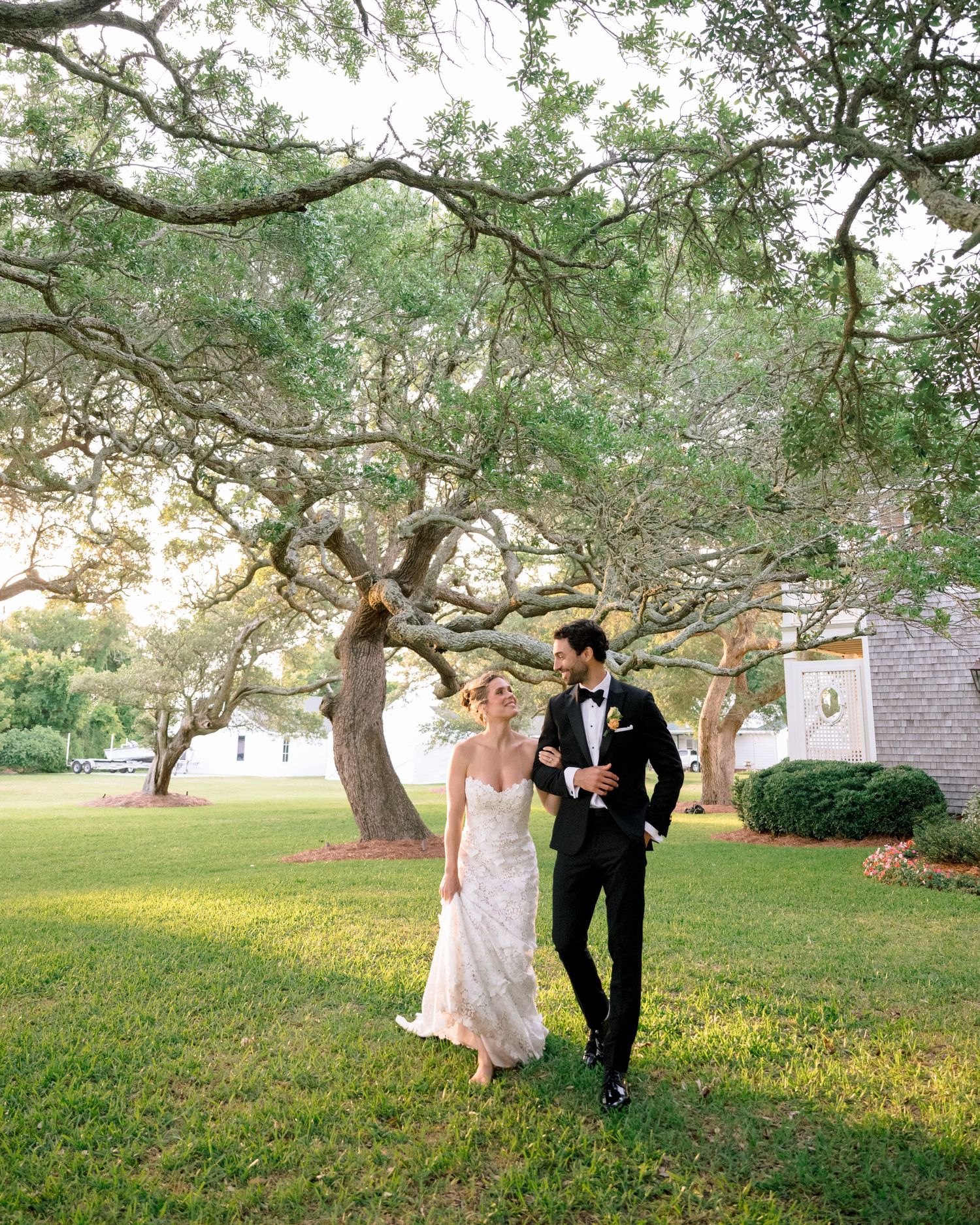 Bride and groom walking hand in hand on lush green lawn beneath sprawling oak trees at an outdoor wedding venue.