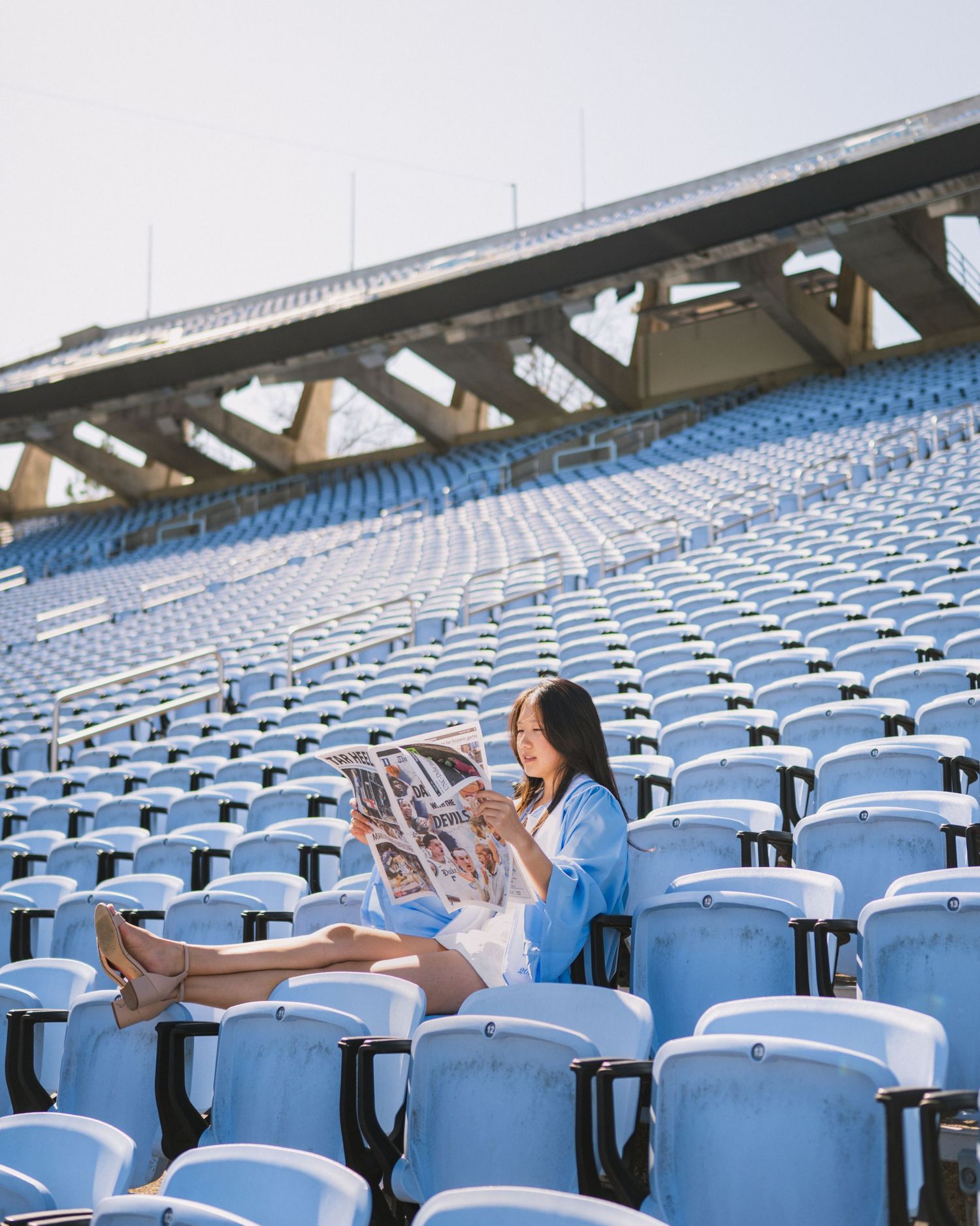 Someone in a blue outfit sits alone in an empty stadium with rows of light blue seats while reading a newspaper.