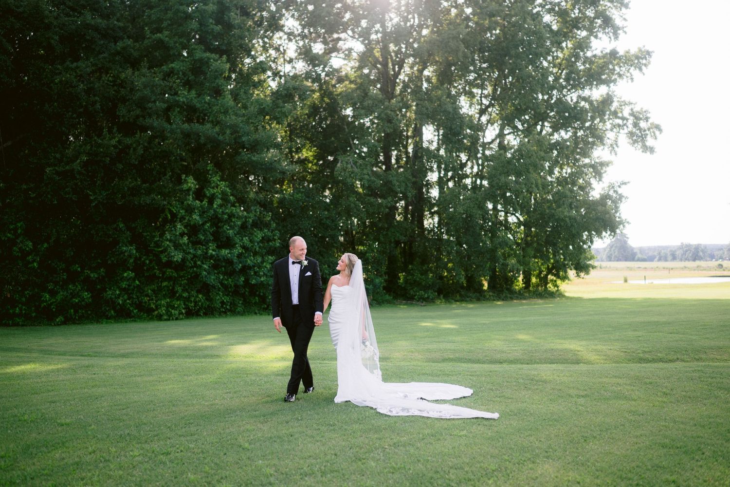 Bride and groom walk hand-in-hand across lush green golf course lawn with tall trees and golden sunlight in background.