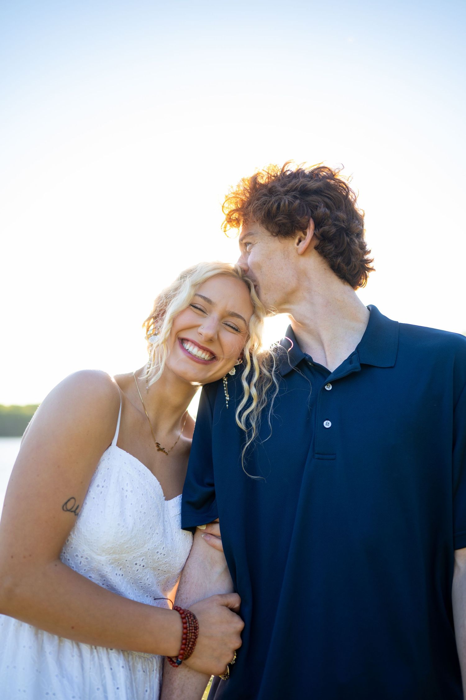 A happy couple embraces outdoors during golden hour, with one wearing a white dress and the other in a navy polo.