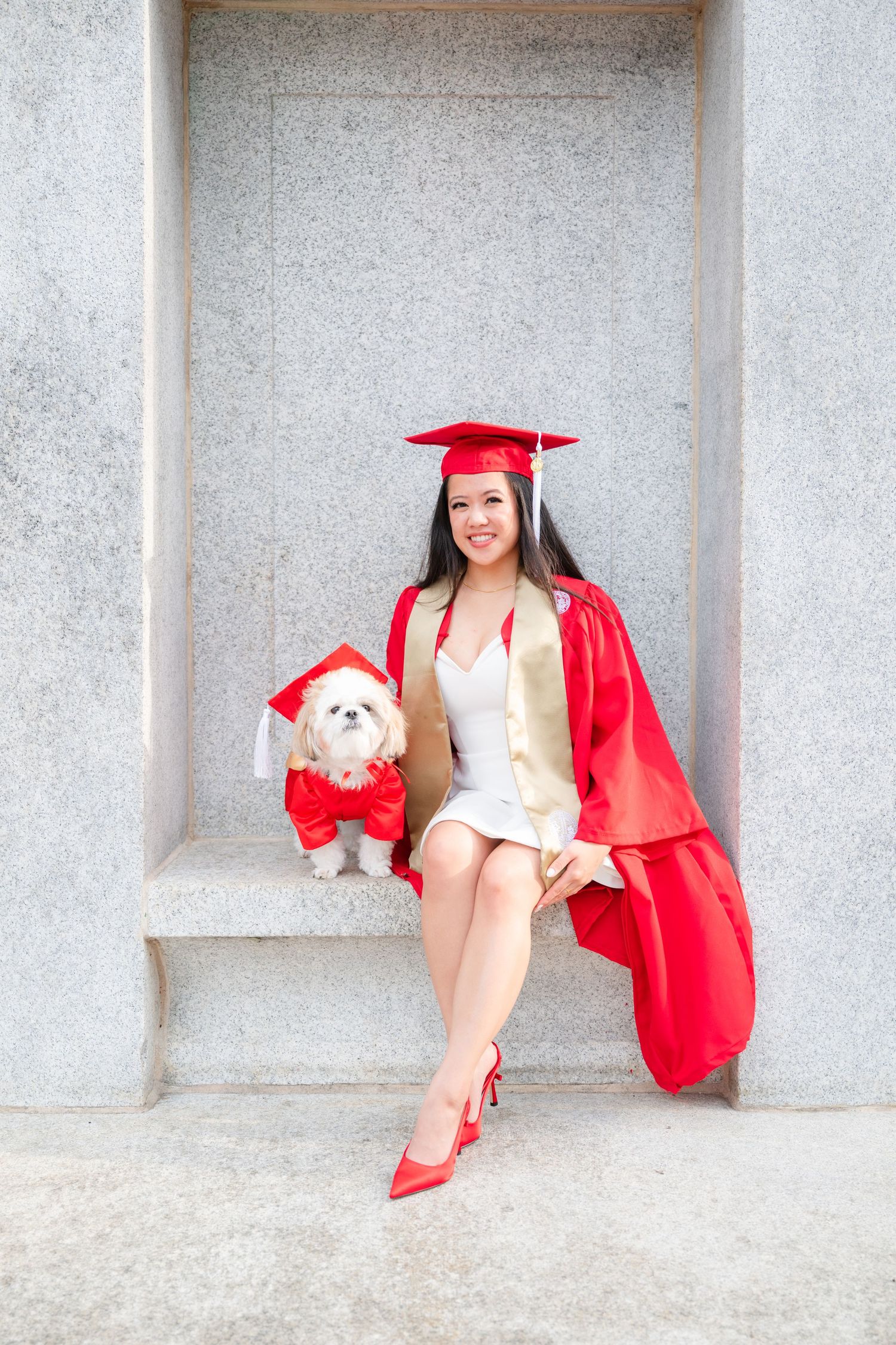Graduate in red cap and gown sits with matching-dressed small dog during graduation photo session.