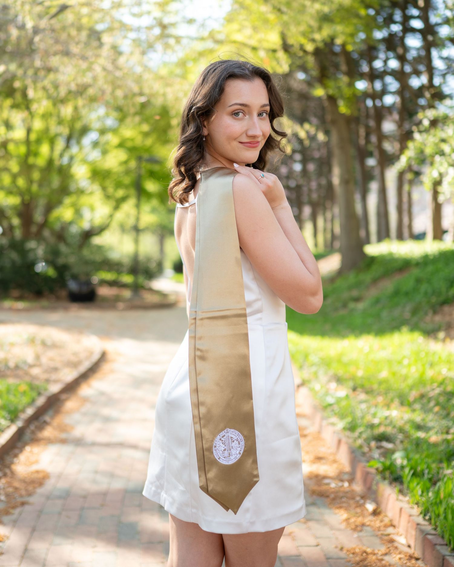 A woman in a white sleeveless top and tan backpack walks along a tree-lined brick pathway on a sunny day.