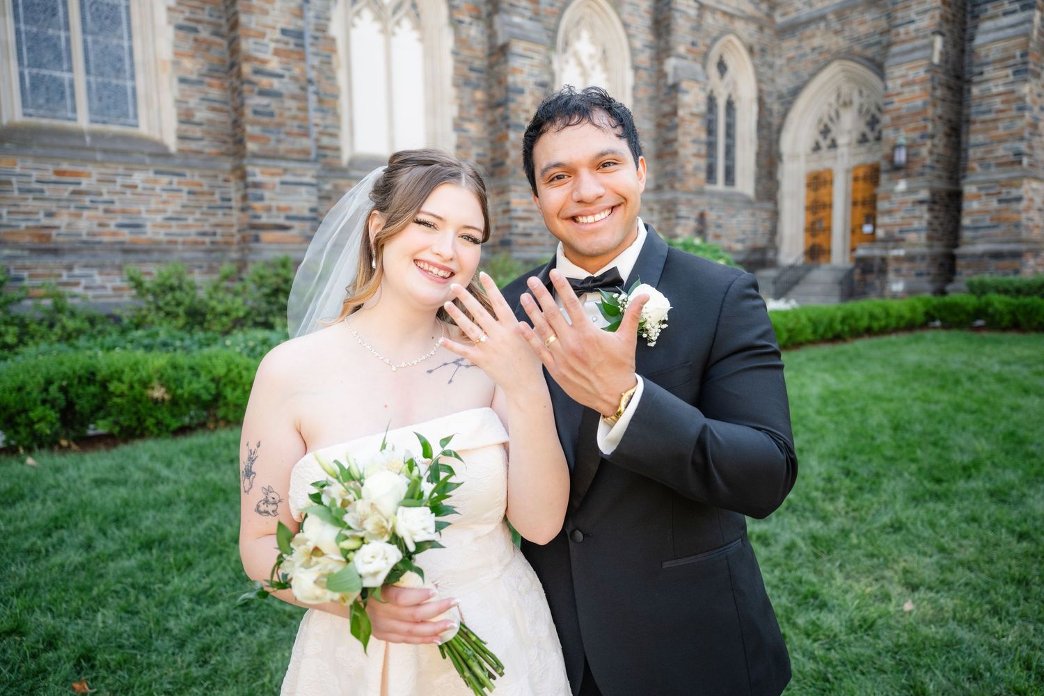 Bride and groom smiling and showing wedding rings in front of a stone church, bride holding white bouquet.