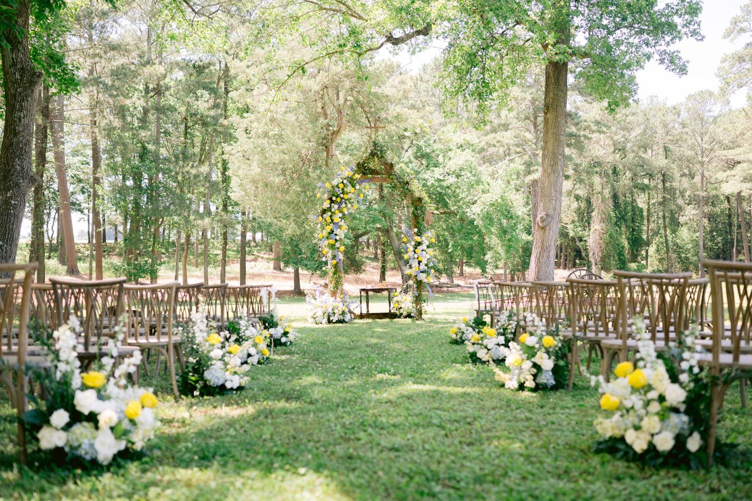 Outdoor wedding ceremony setup with wooden chairs, floral arch, and yellow and white flowers lining the aisle in a sunlit forest.