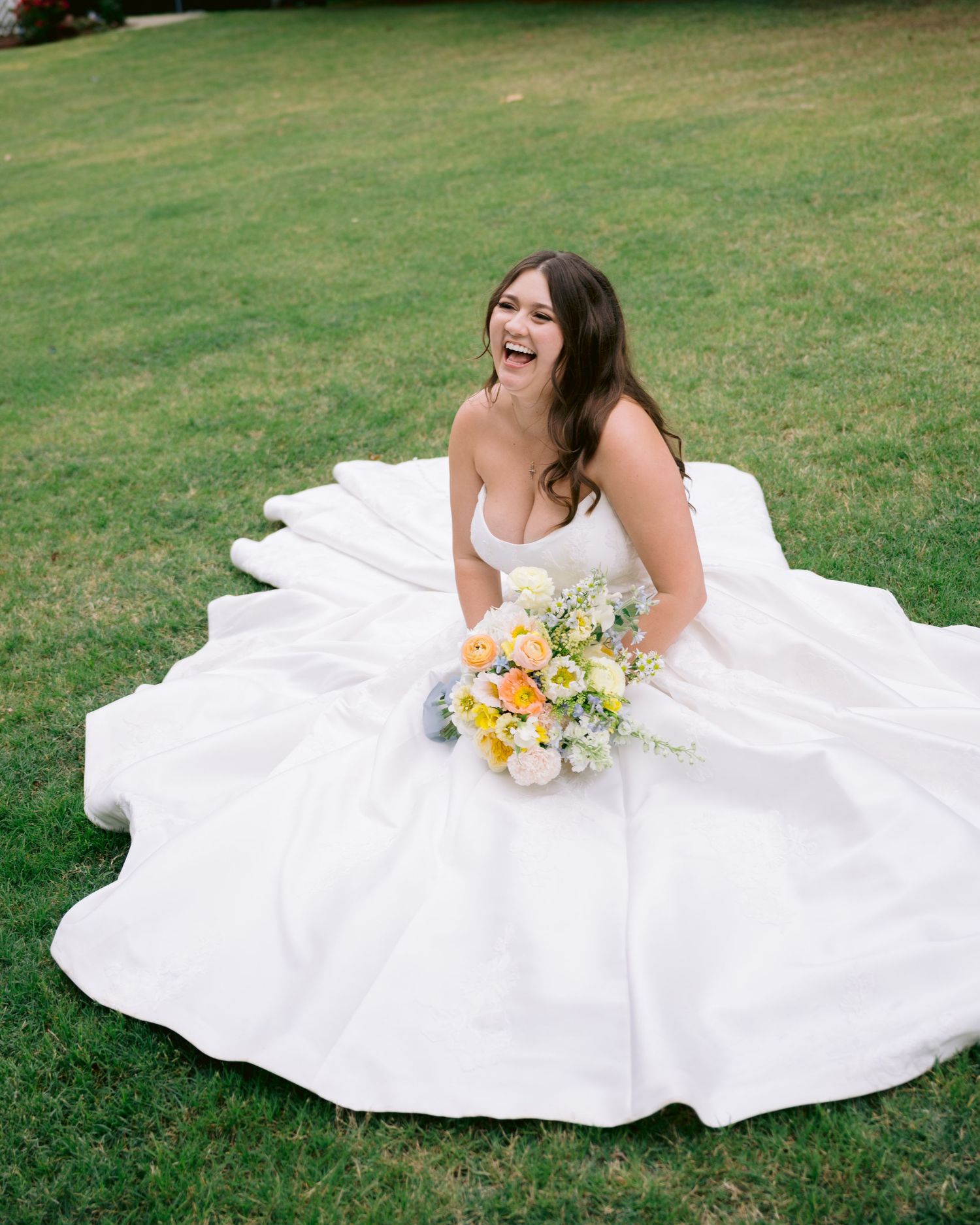 Laughing bride in a white ballgown sits on green grass holding a colorful bouquet of yellow, orange, and white flowers.
