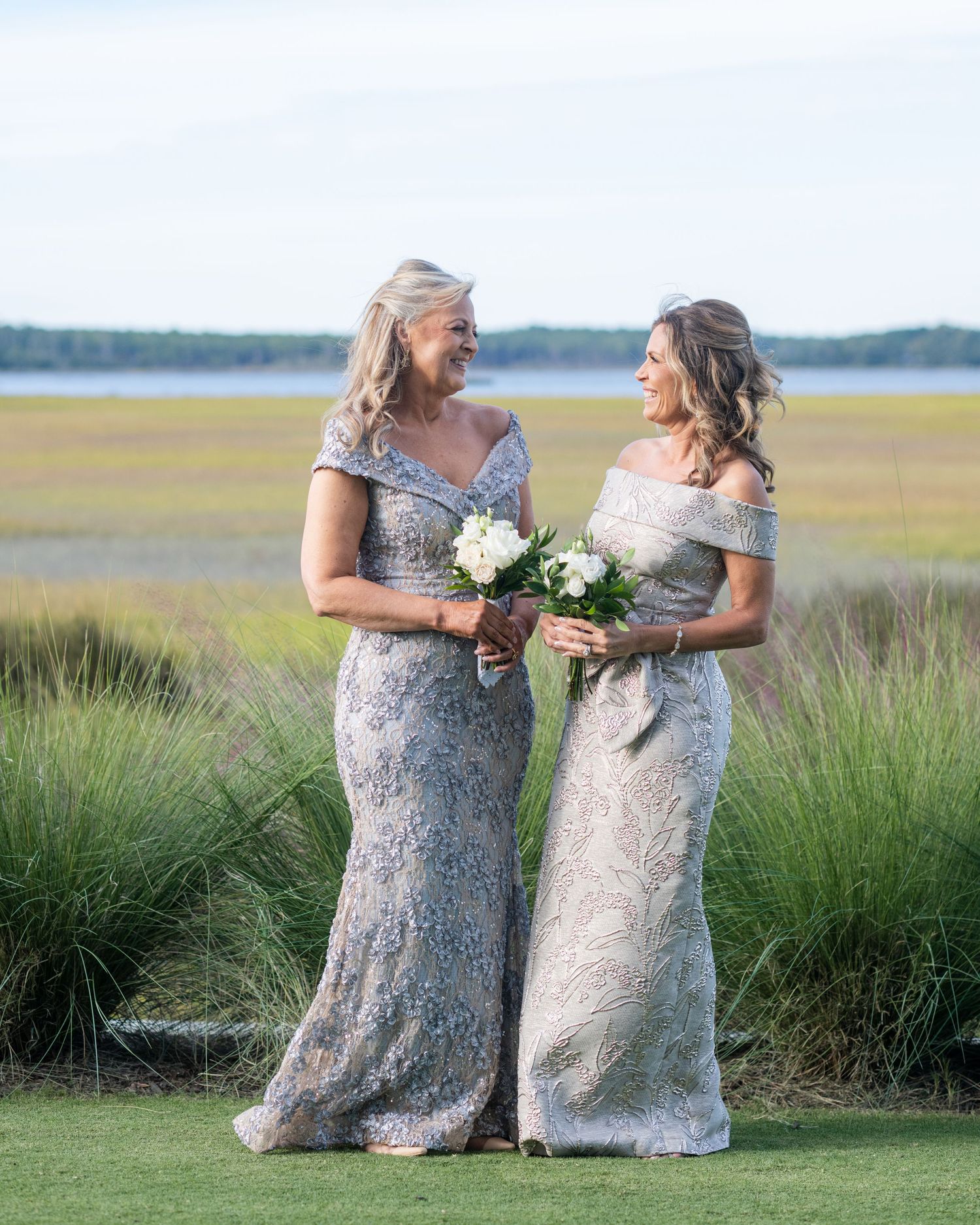 Two bridesmaids in silver sequined dresses share a joyful moment together on a grassy coastal marsh at sunset.