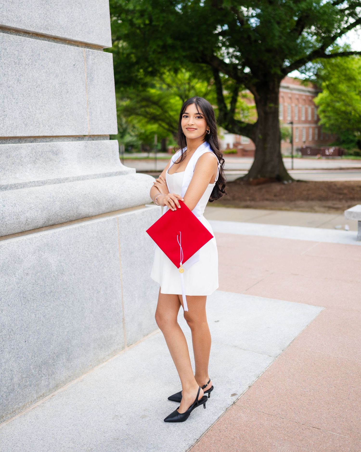 Person in a white dress and black heels poses against a light-colored wall outdoors on a sunny day.