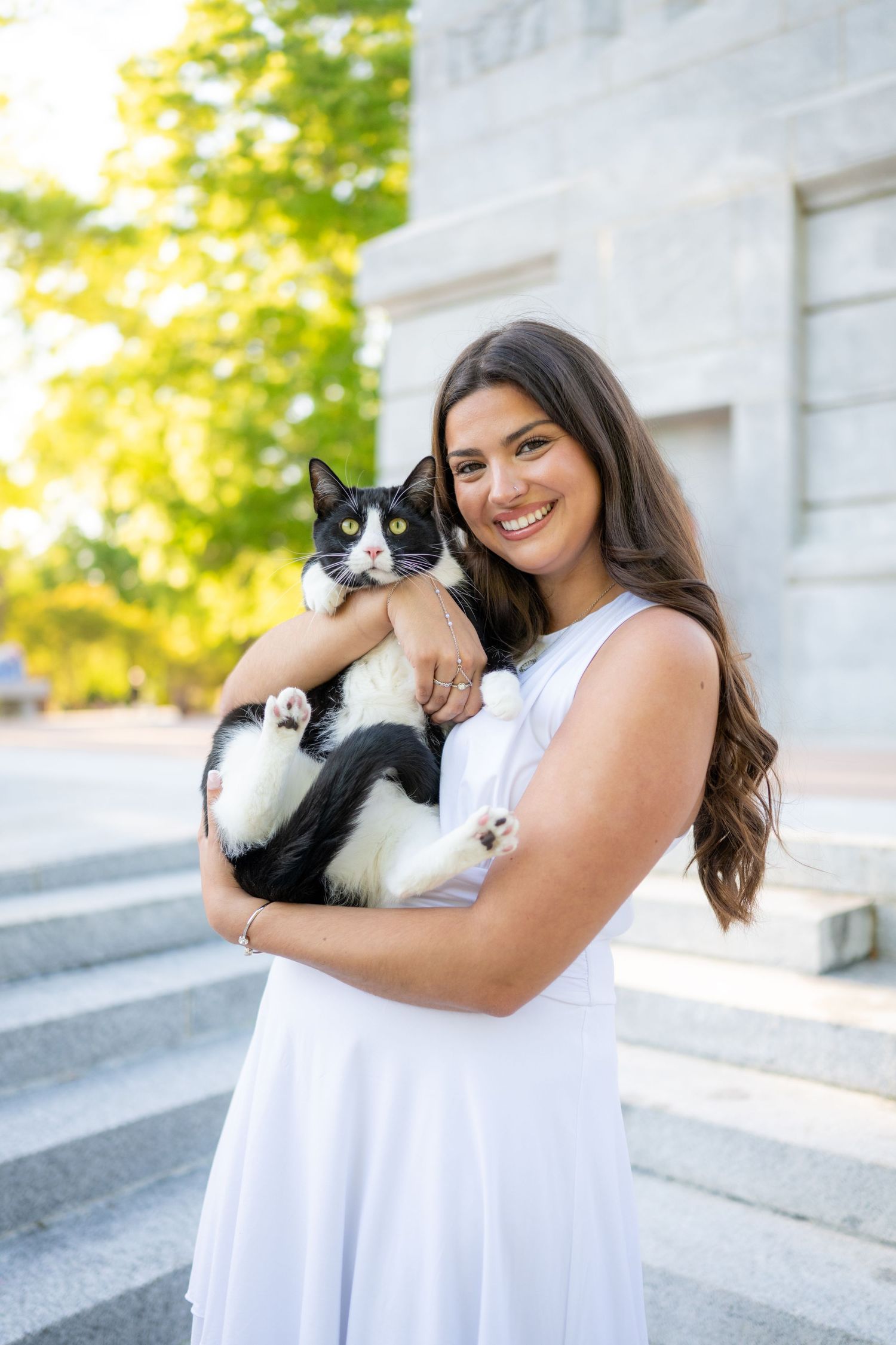 A person in a white dress holds a black and white cat outdoors on a sunny day near stone steps.
