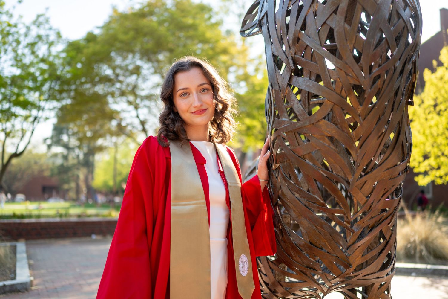 A person in a bright red blazer and white top poses next to a decorative metal sculpture in an outdoor setting.