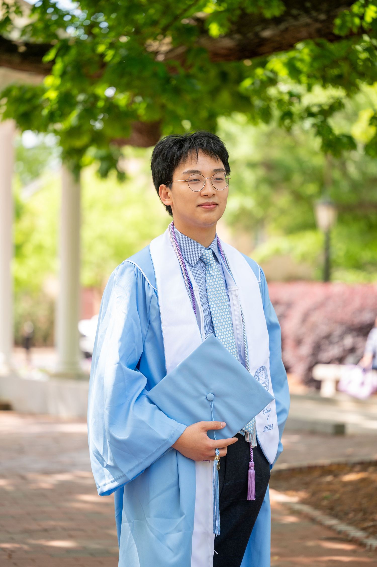 A graduate in light blue academic regalia stands outdoors under green foliage holding a folder on a sunny day.