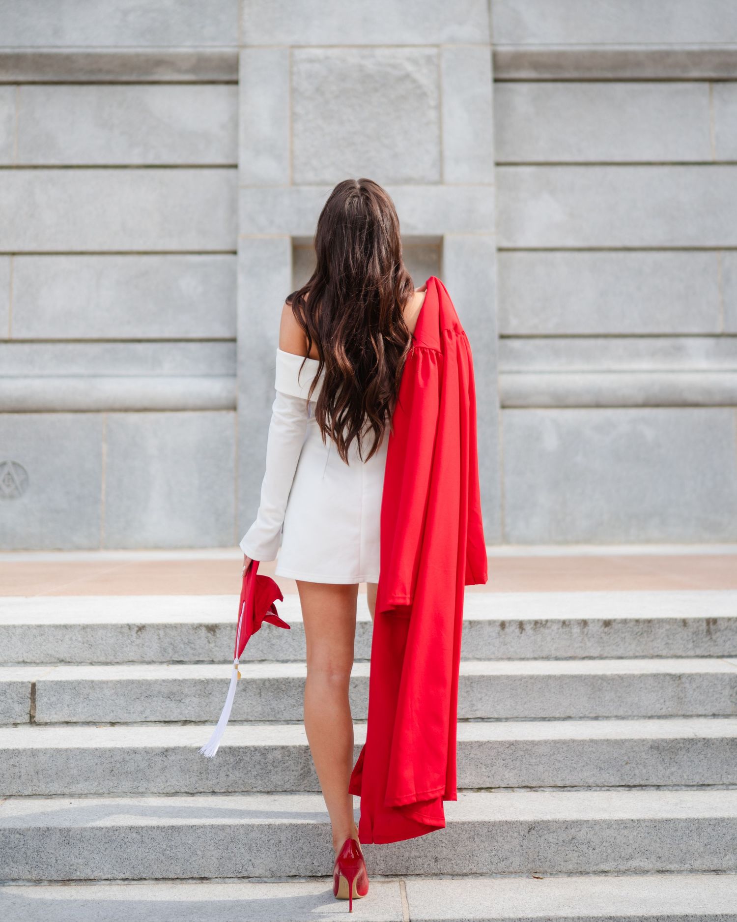 A person in white clothing with a flowing red fabric descends stone steps against a light stone wall backdrop.