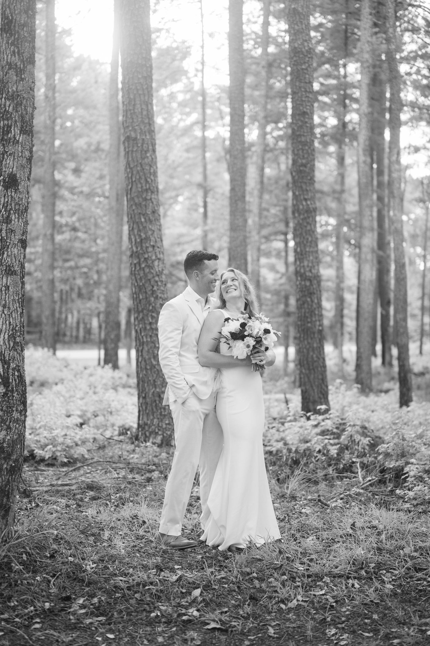 A romantic black and white wedding photo series showing a couple in white attire embracing in a misty pine forest setting.