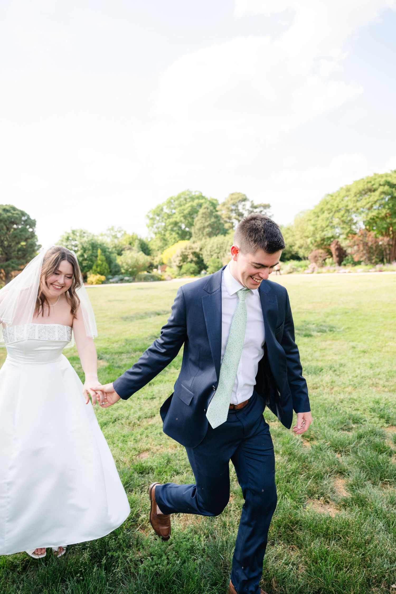 Joyful bride and groom holding hands running across green lawn in wedding attire on sunny day.