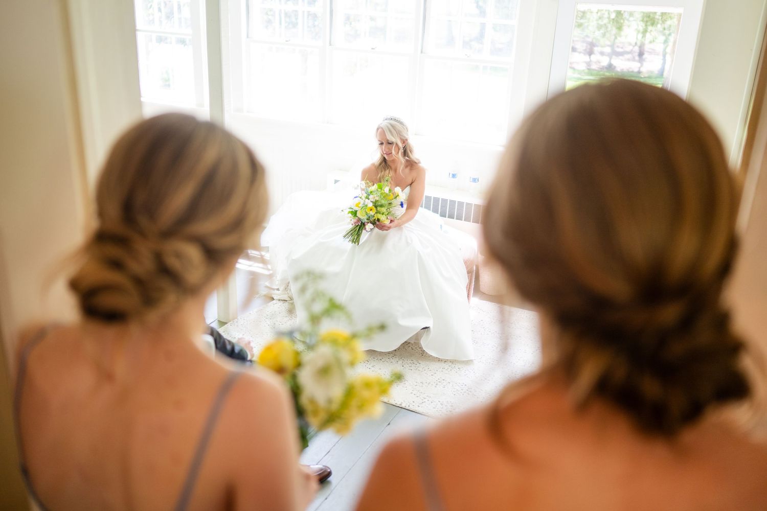 A bride and her bridesmaids in peach dresses stand in a row looking out a window during wedding preparations.