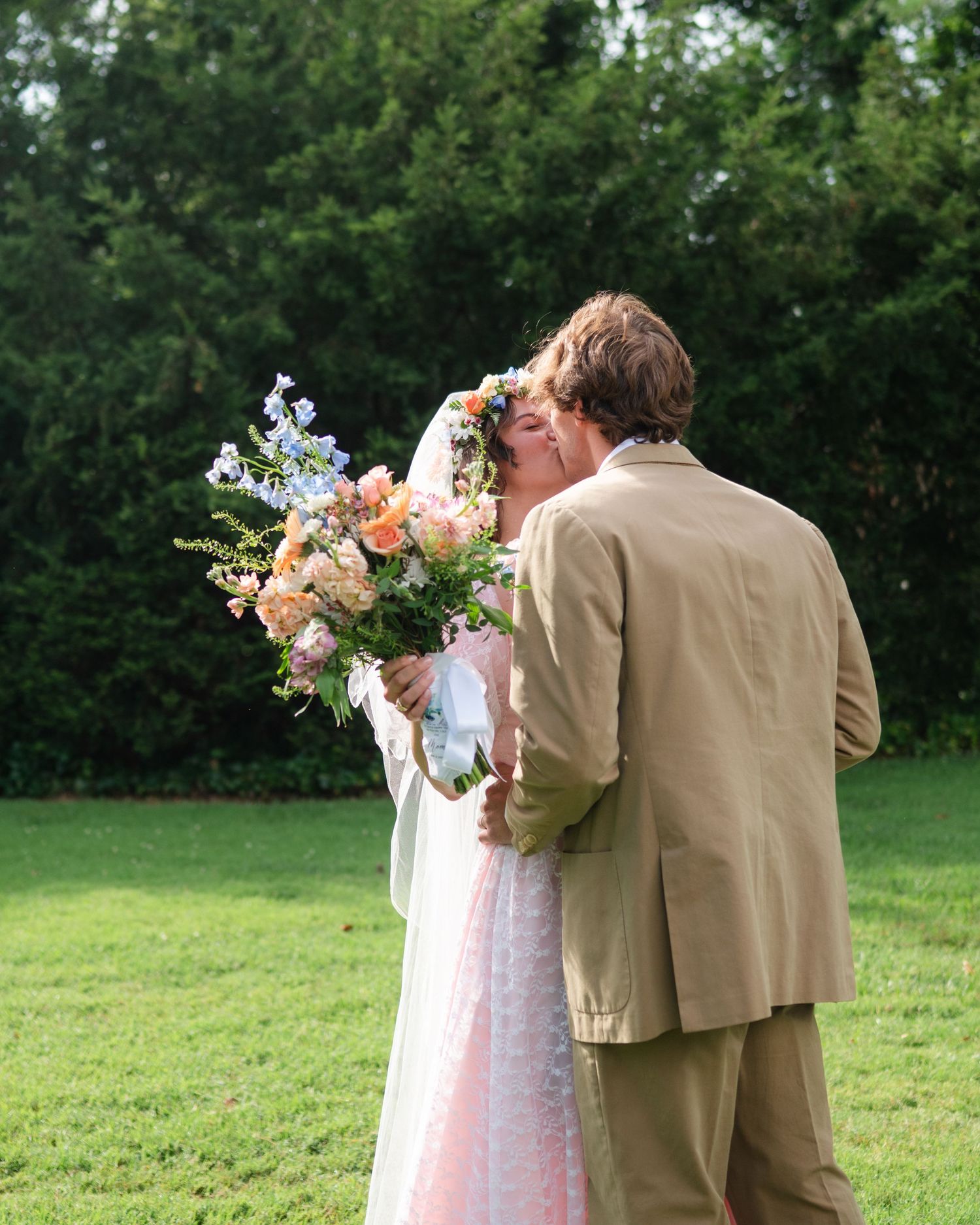A couple in wedding attire stands in a green field, with the bride holding a colorful floral bouquet.