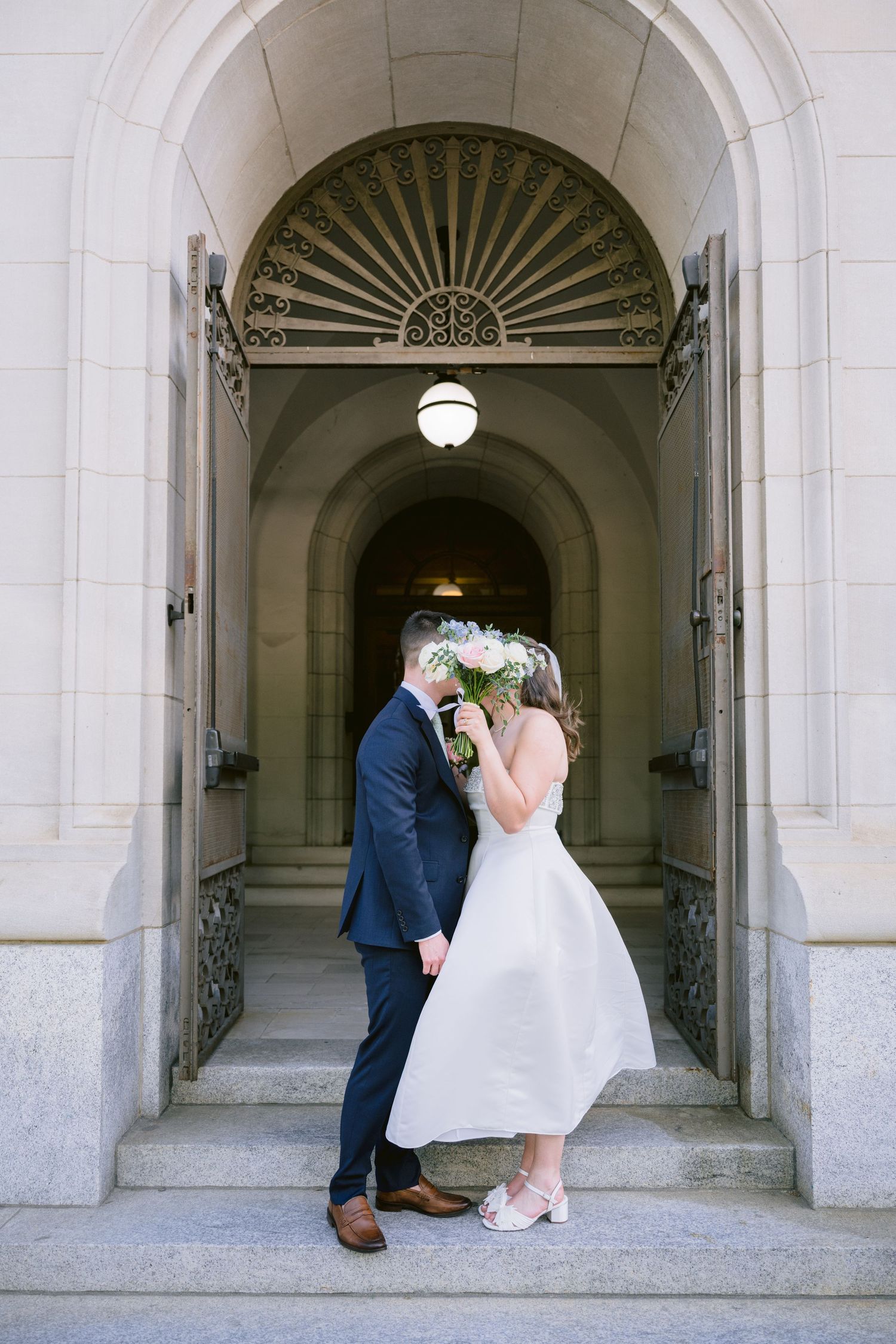 Bride and groom kissing behind bouquet in elegant arched doorway of stone building on wedding day.