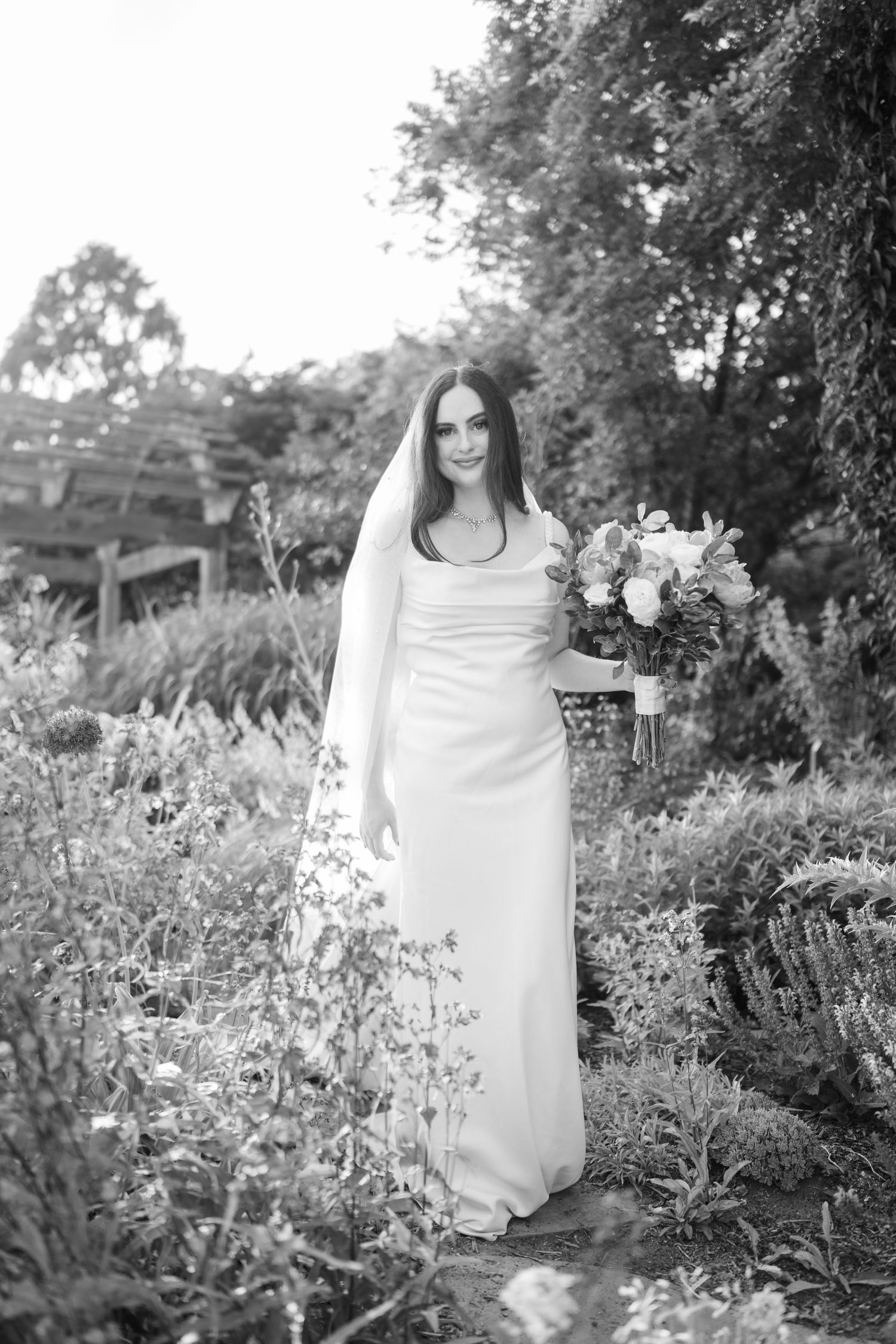 Bride in white gown and veil holds bouquet in lush garden, black and white bridal portrait photography.