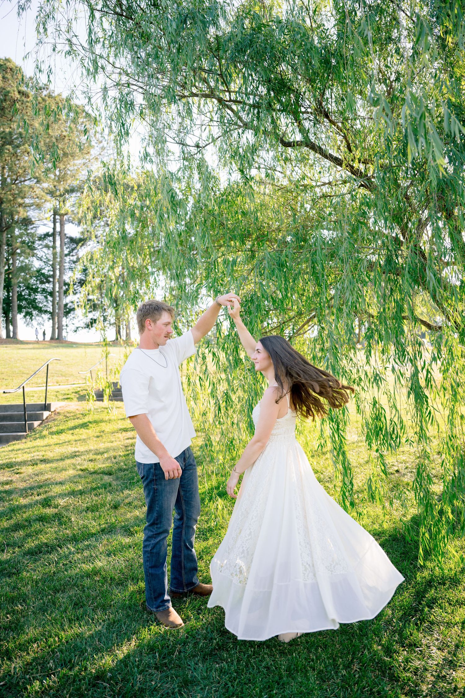 Couple dancing under a weeping willow tree on green grass, woman in white dress twirling, man in white shirt and jeans.