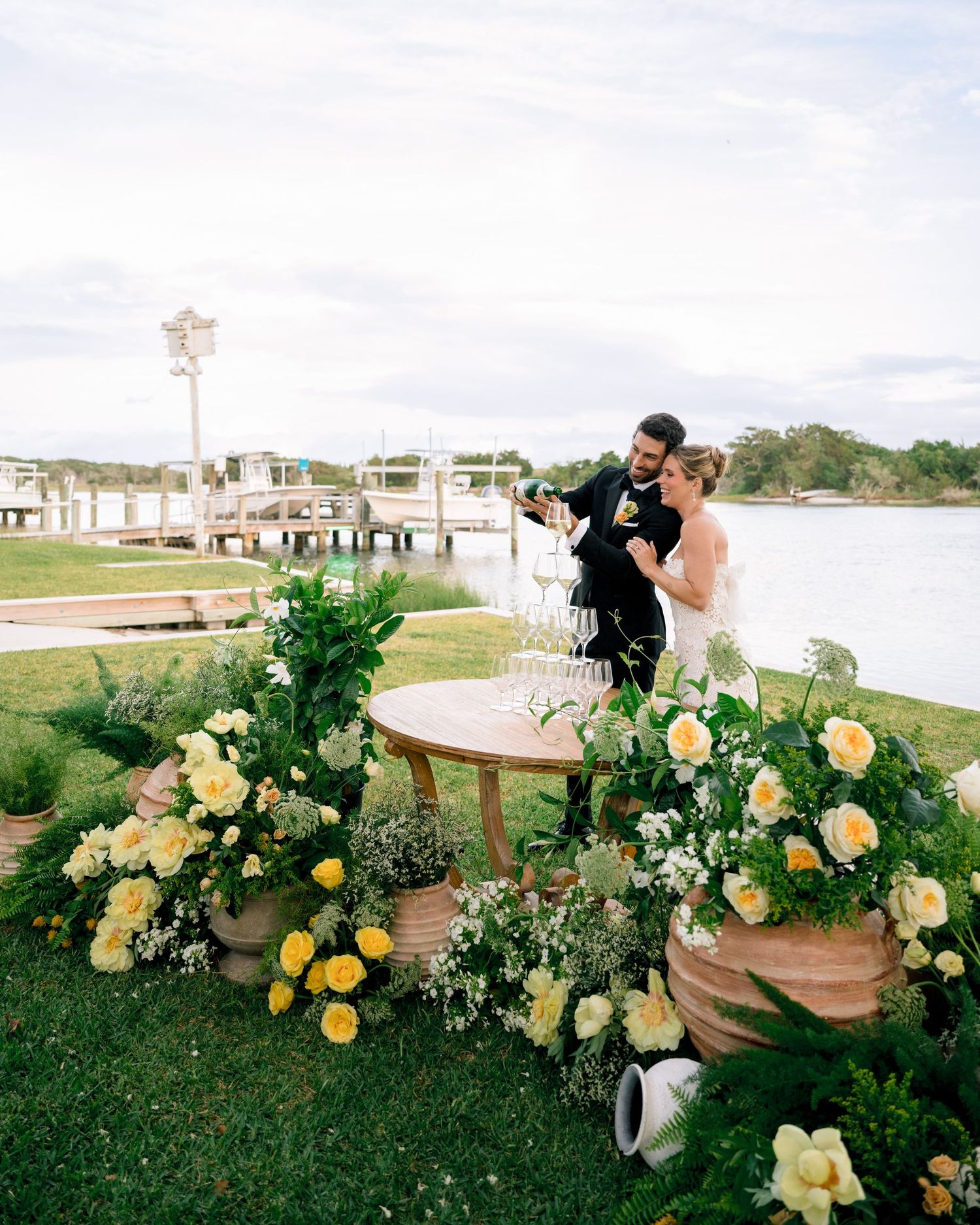 Bride and groom pouring champagne tower surrounded by yellow floral arrangements beside a waterfront venue.