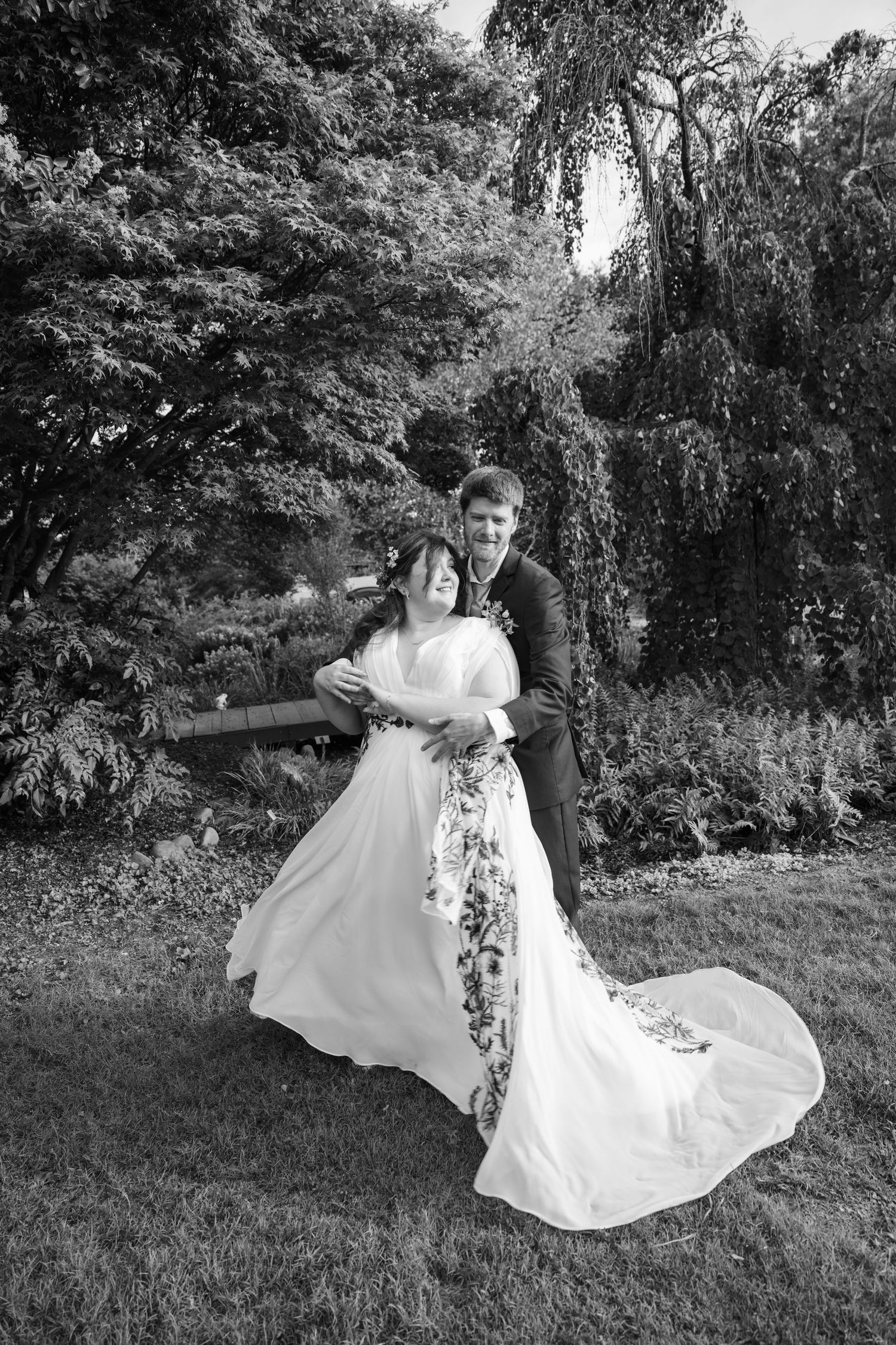 Black and white photo of a bride and groom dancing outdoors in a lush garden, her floral wedding dress train flowing gracefully.