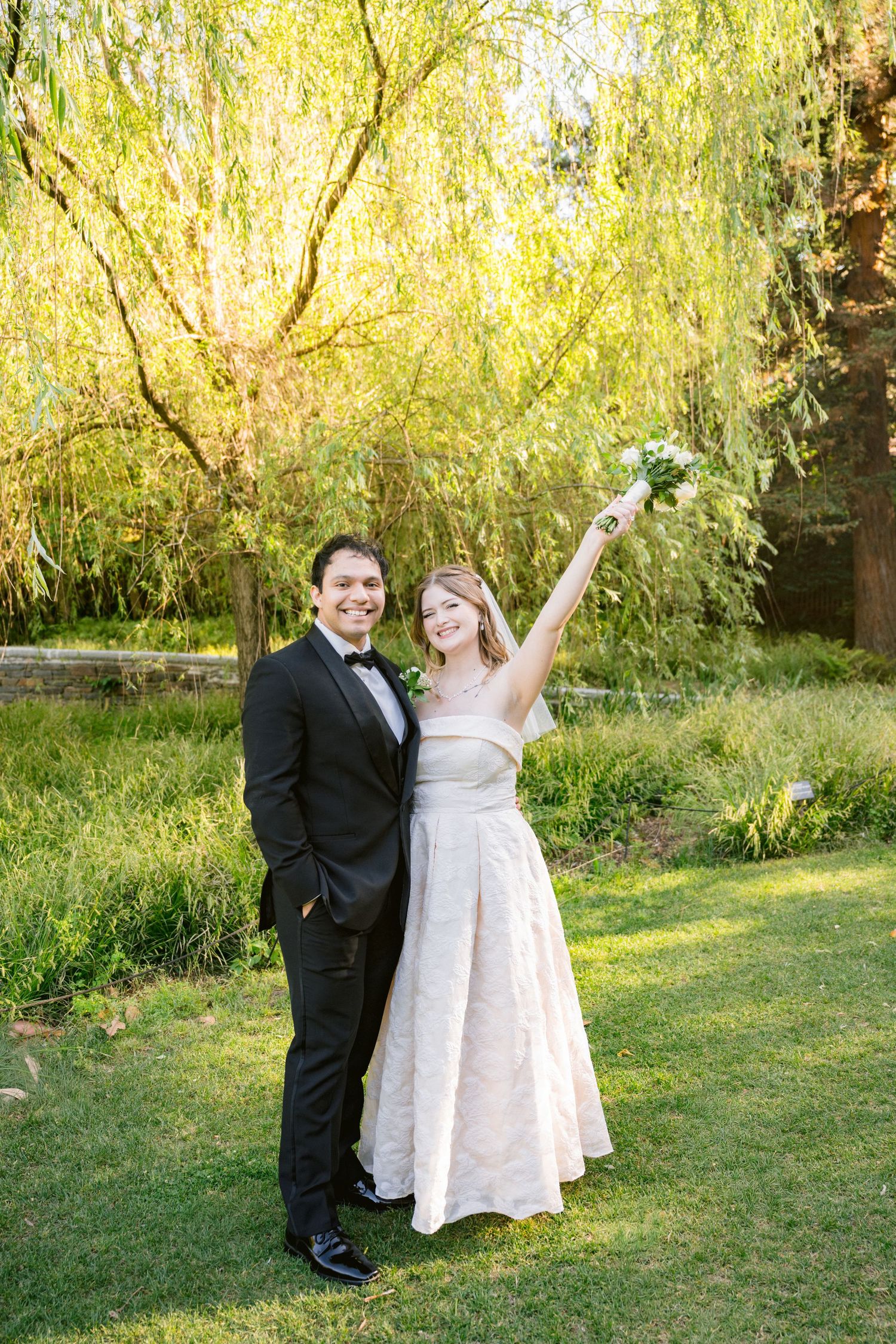 Joyful bride in white gown raising bouquet beside groom in black tuxedo, standing on green lawn under willow tree.