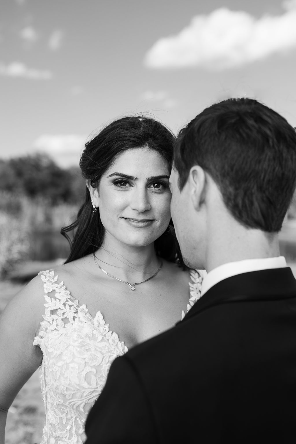 Black and white portrait of smiling bride in lace gown gazing at groom under blue sky with clouds.
