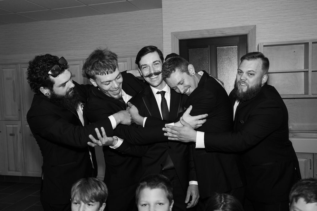Groom surrounded by groomsmen in black suits laughing and playfully hugging in a black and white photo.