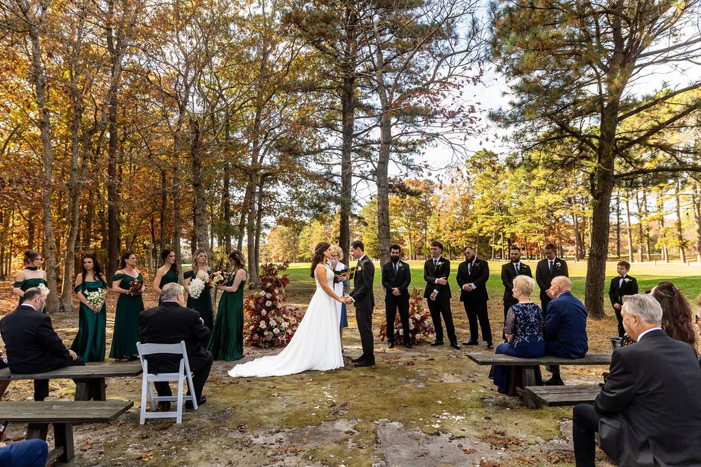 Outdoor autumn wedding ceremony in wooded setting with wedding party and seated guests among fall foliage.