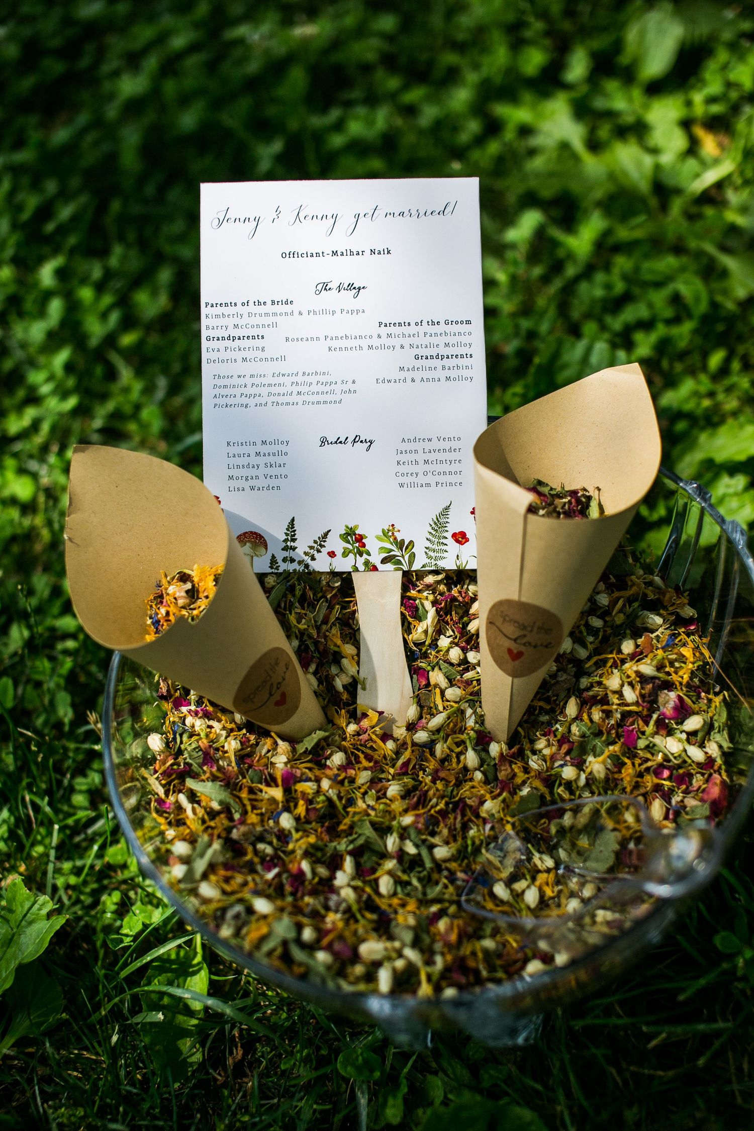 Wedding program and paper cones filled with flower petals on grass.